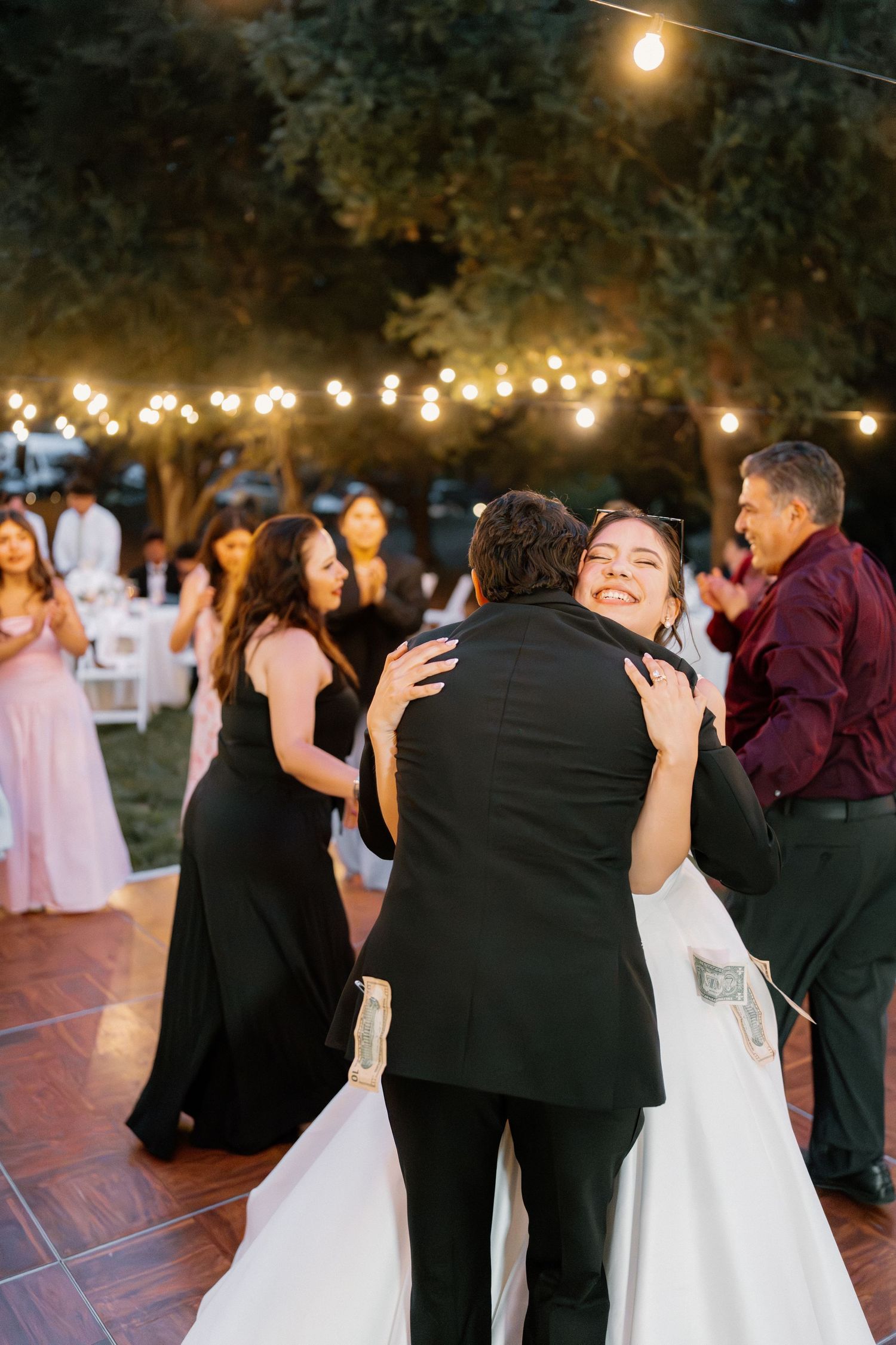 Smiling bride embraces groom on outdoor dance floor surrounded by guests and glowing string lights at night.