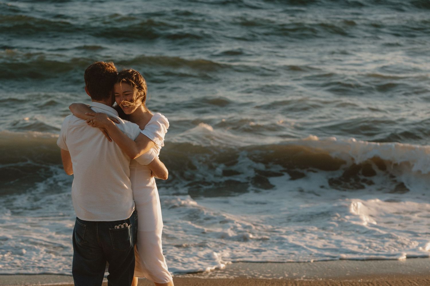 Romantic couple hugging on a beach at sunset with gentle waves crashing behind them.