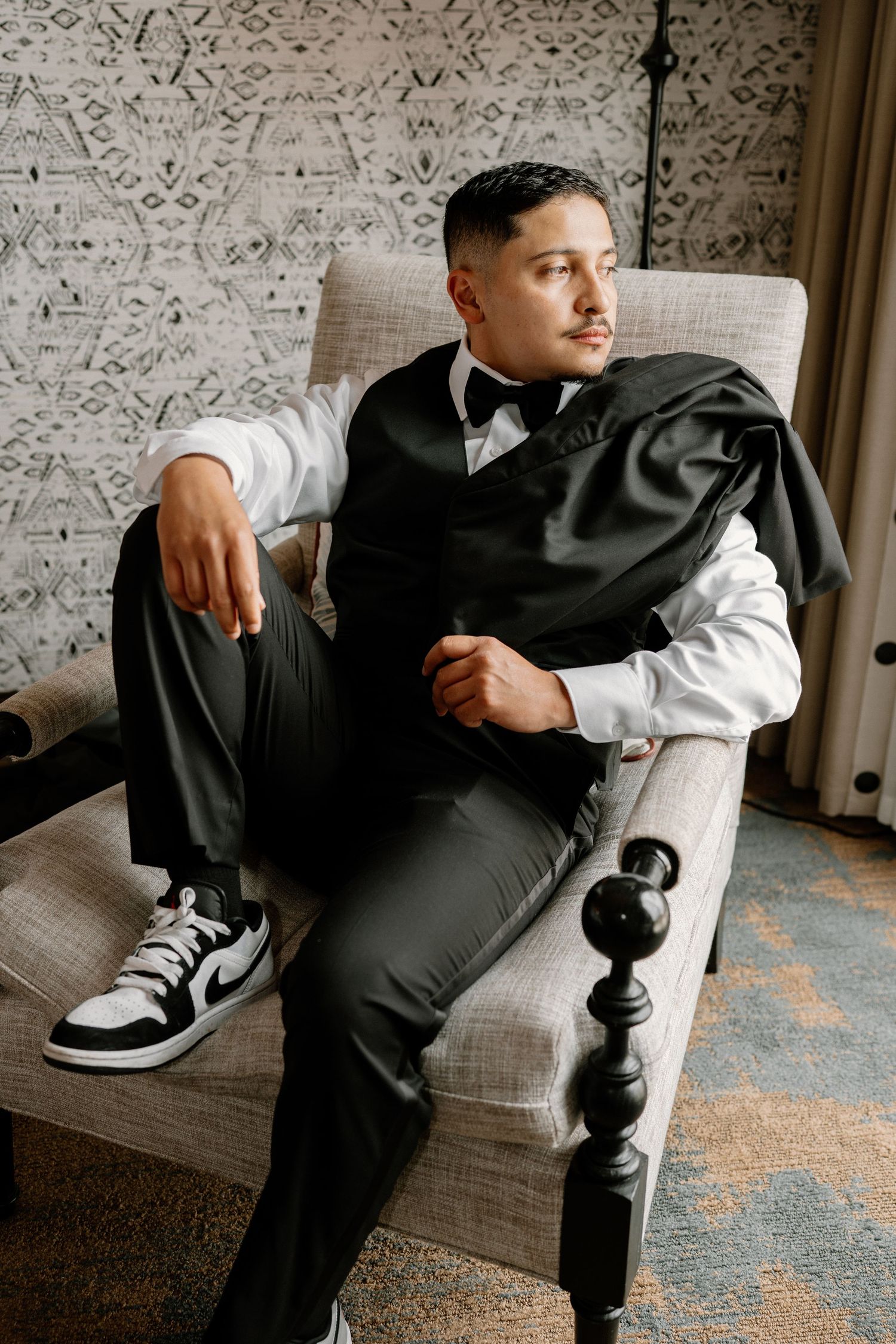 Groom in black tuxedo and bow tie sitting in armchair wearing Nike Air Jordan sneakers before wedding.
