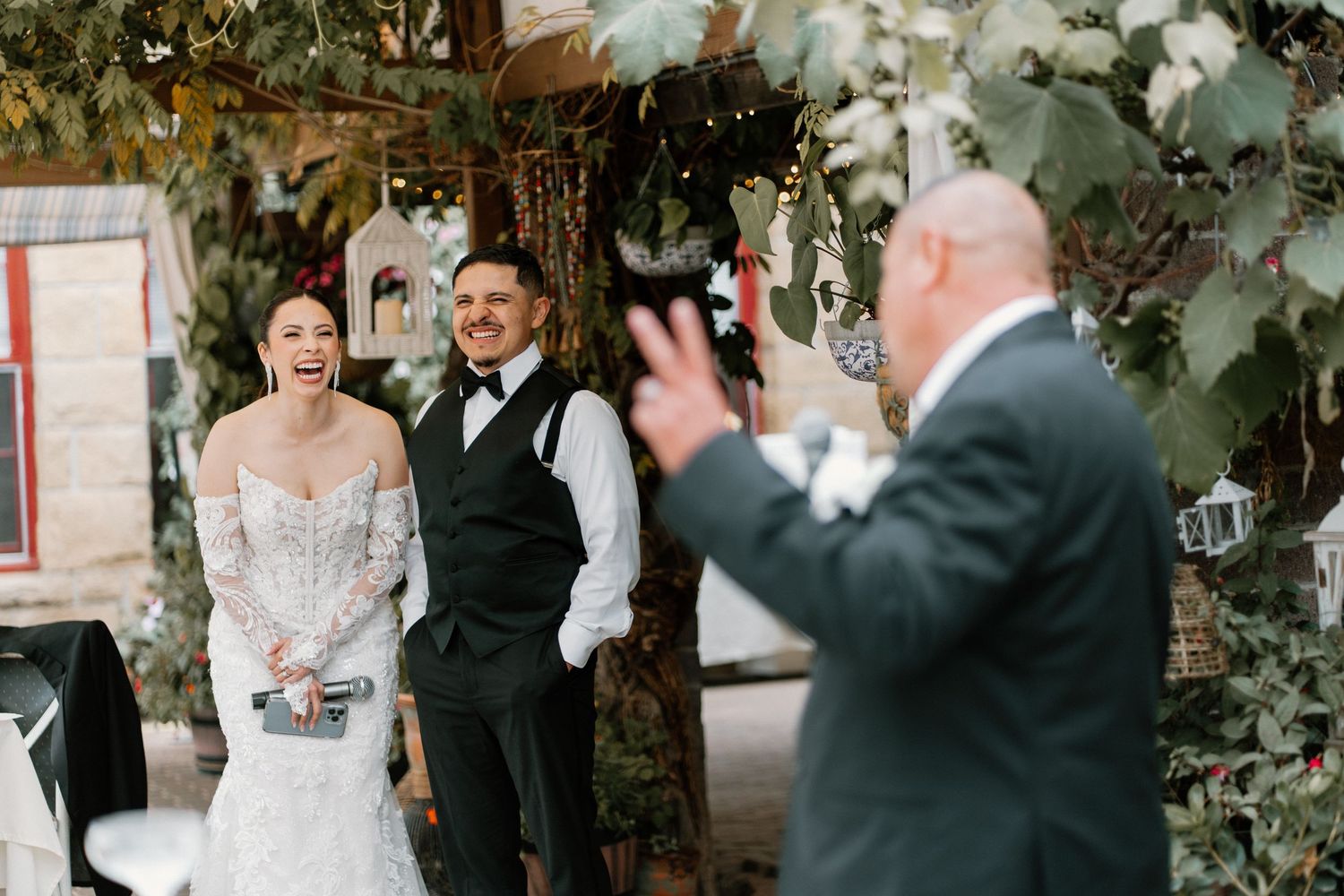 Bride and groom laughing joyfully with guest at rustic outdoor wedding reception with string lights.