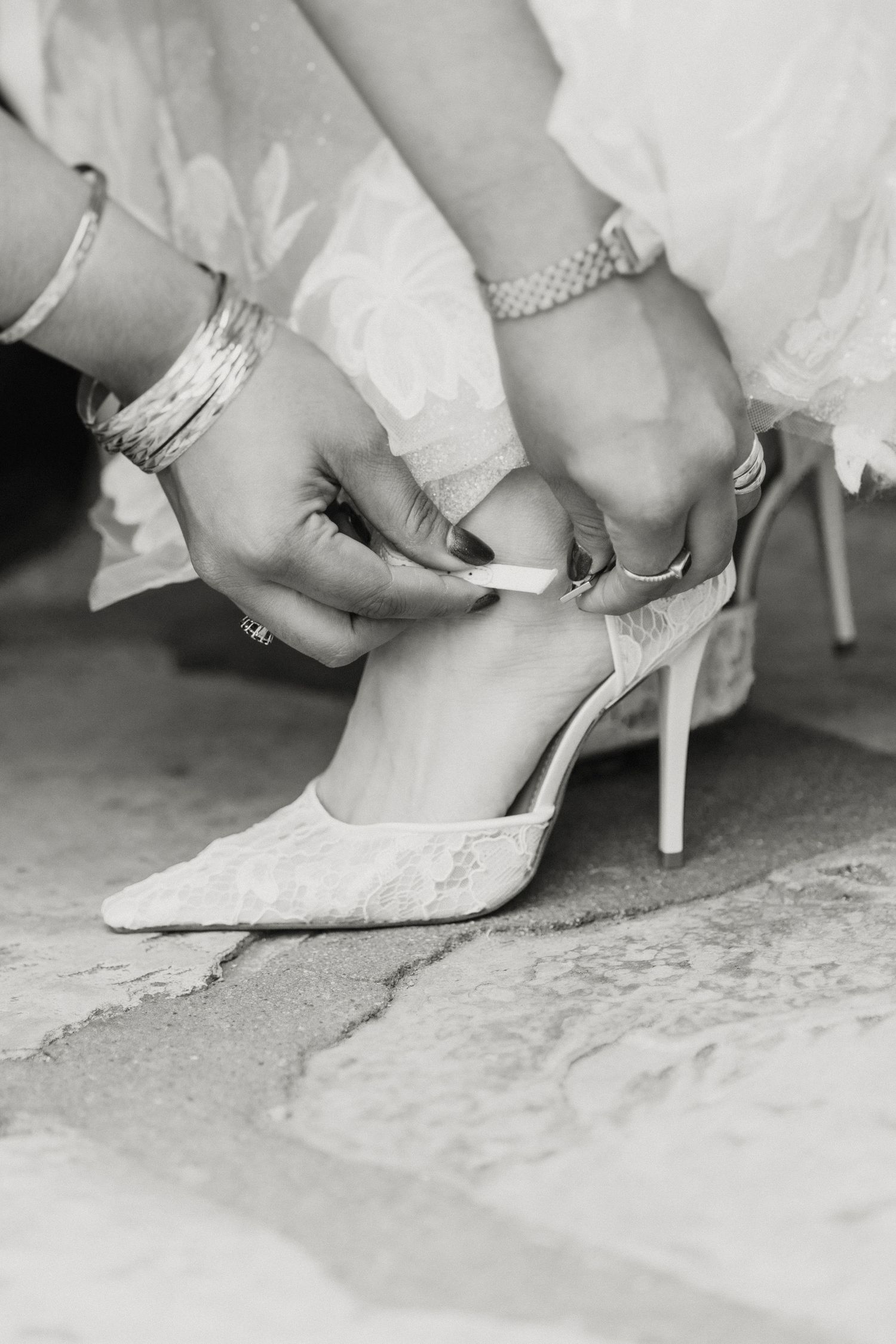 Black and white close-up of hands fastening the strap of a lace bridal high heel wedding shoe.