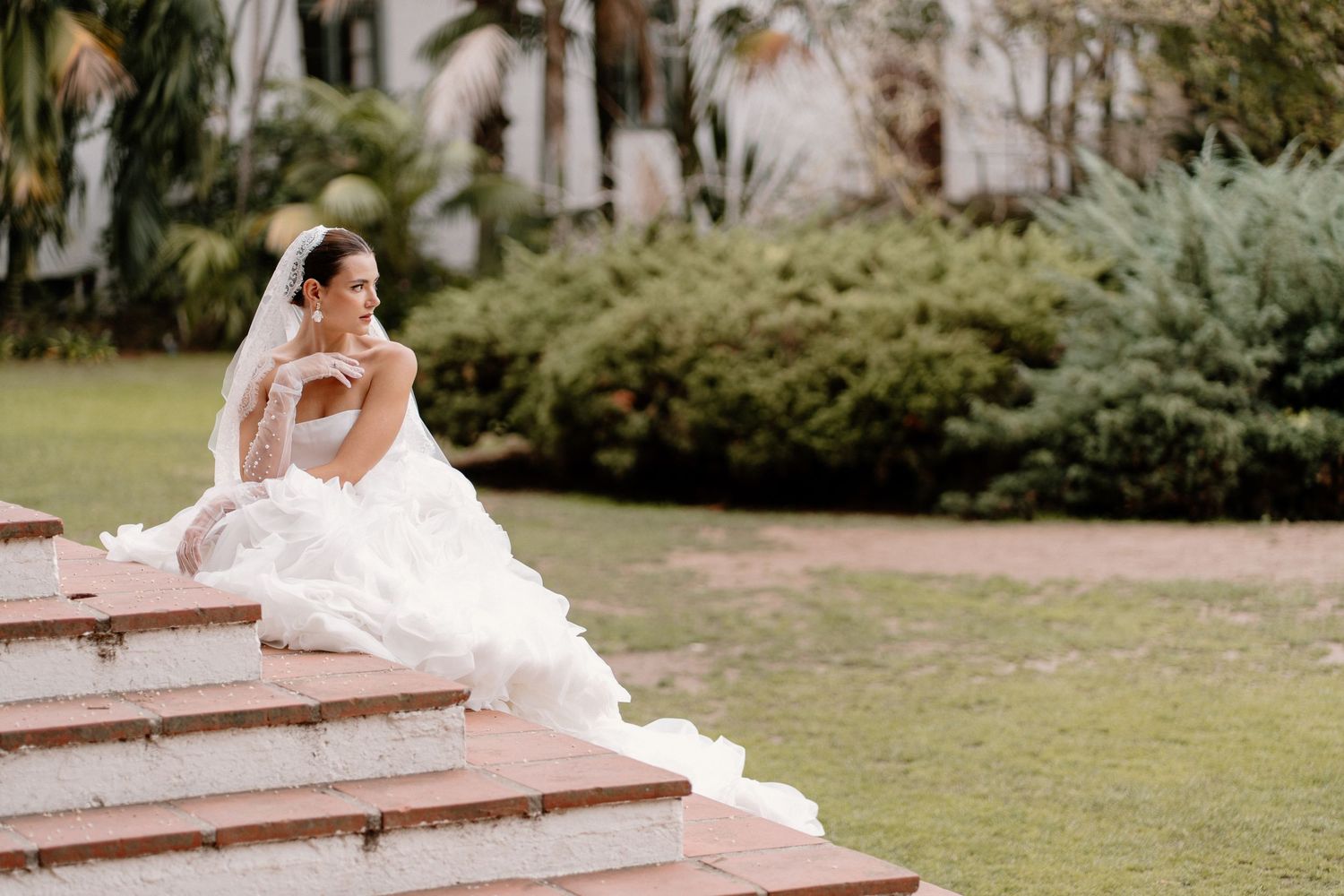 Bride in voluminous white gown and veil sitting gracefully on outdoor stone steps.