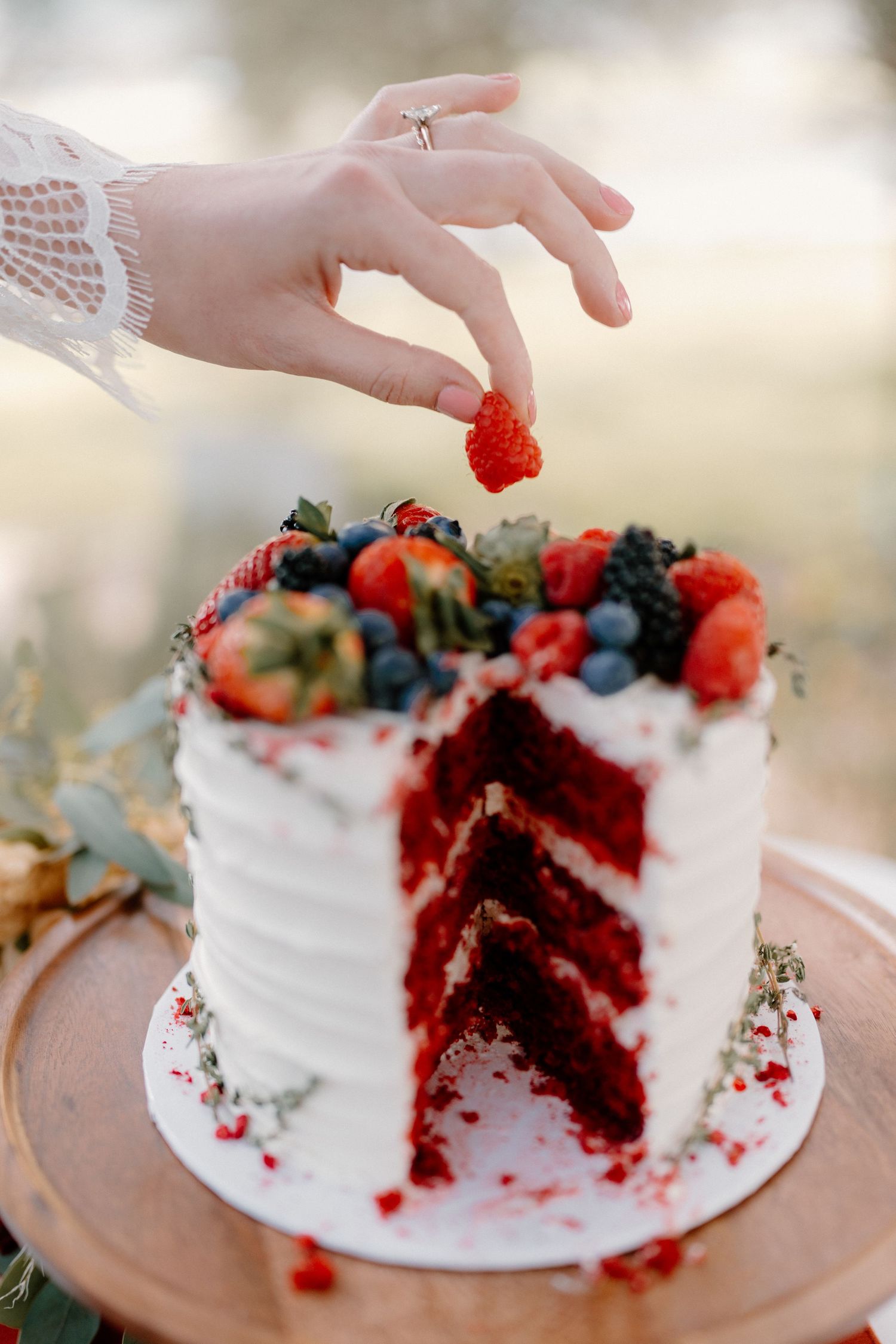 Bride\u2019s hand holding a strawberry over a sliced red velvet wedding cake topped with fresh berries.