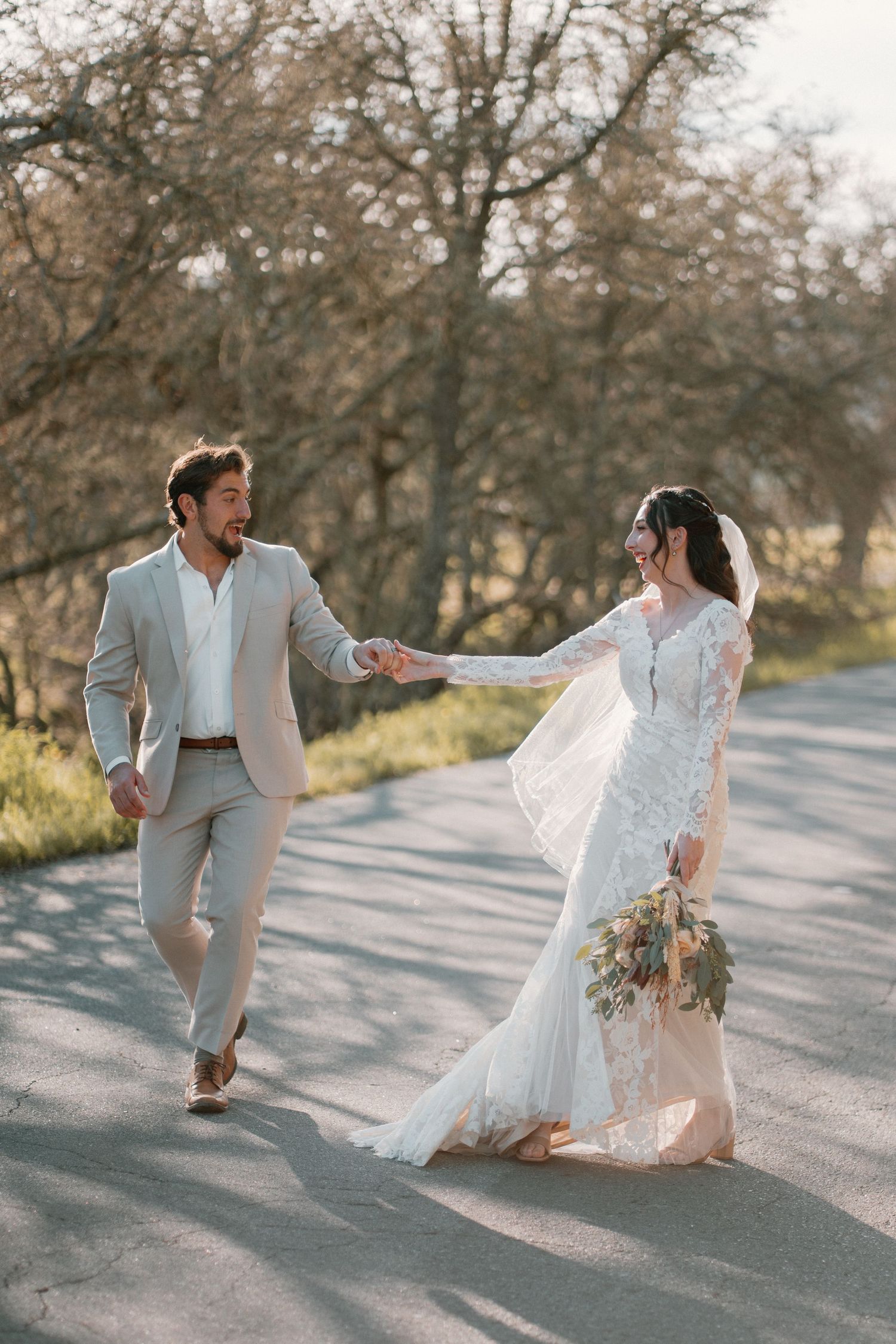 Bride and groom holding hands walking down a sunlit road surrounded by bare trees in wedding attire.