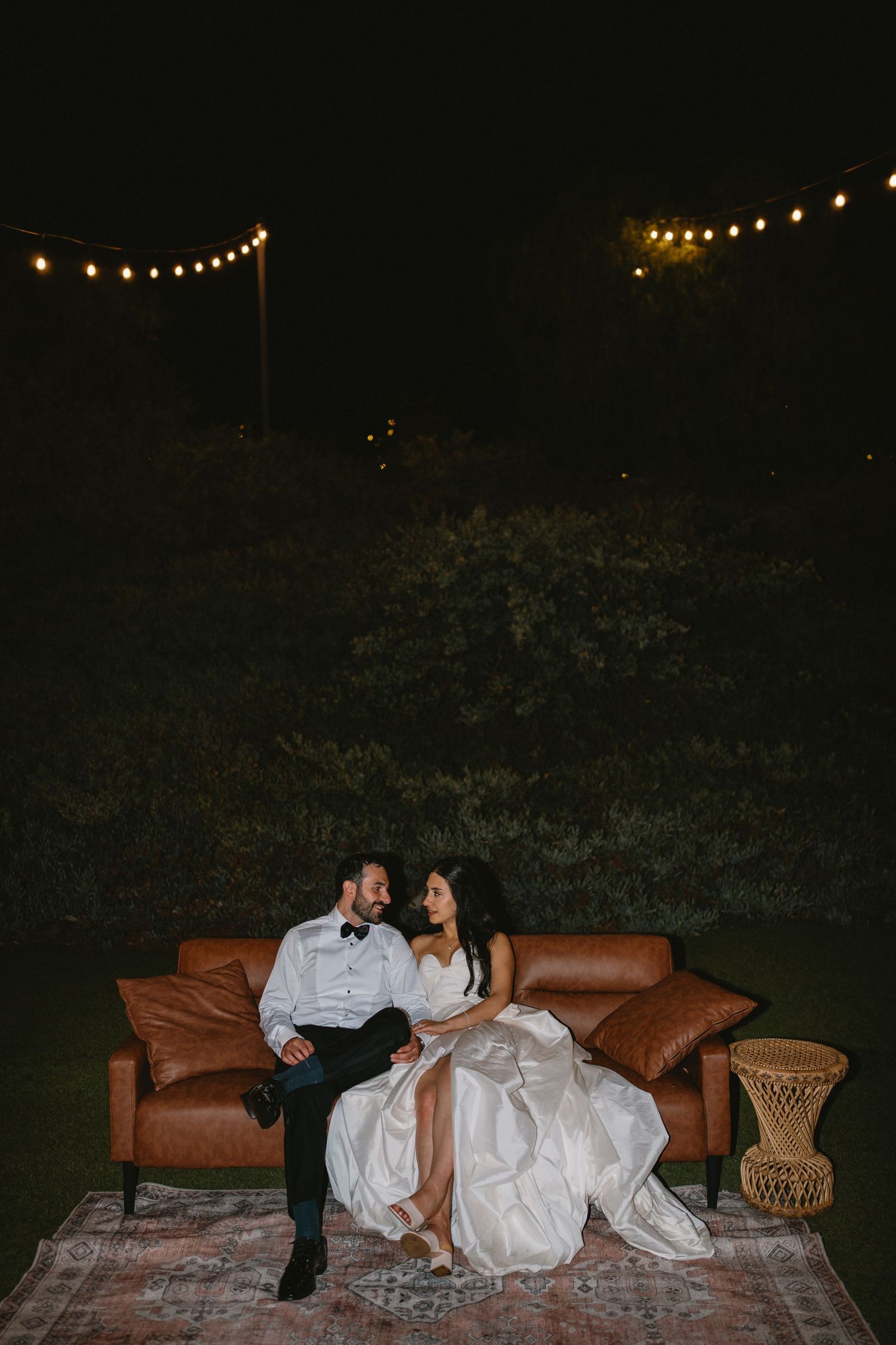 Bride and groom sitting on a brown leather sofa outdoors at night under string lights.