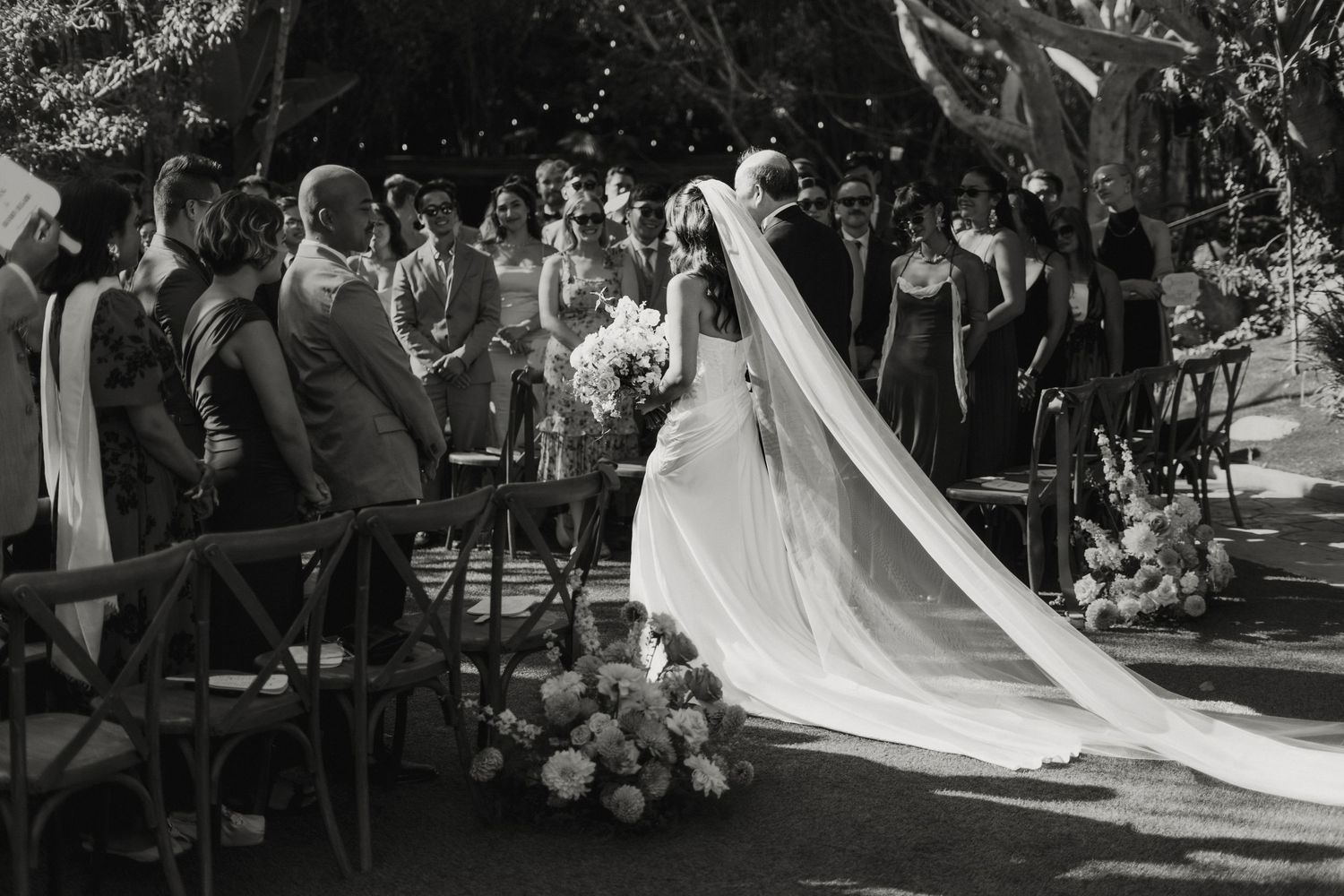 Black and white image of bride walking down aisle in elegant gown and cathedral veil outdoors.