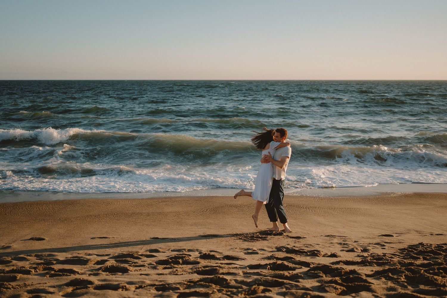 Couple embracing and dancing joyfully on a sandy beach with ocean waves in the background at golden hour.