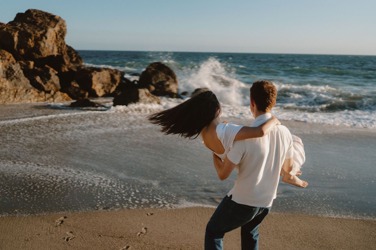 Couple sharing a romantic moment on a rocky beach, man carrying woman as ocean waves crash behind them.