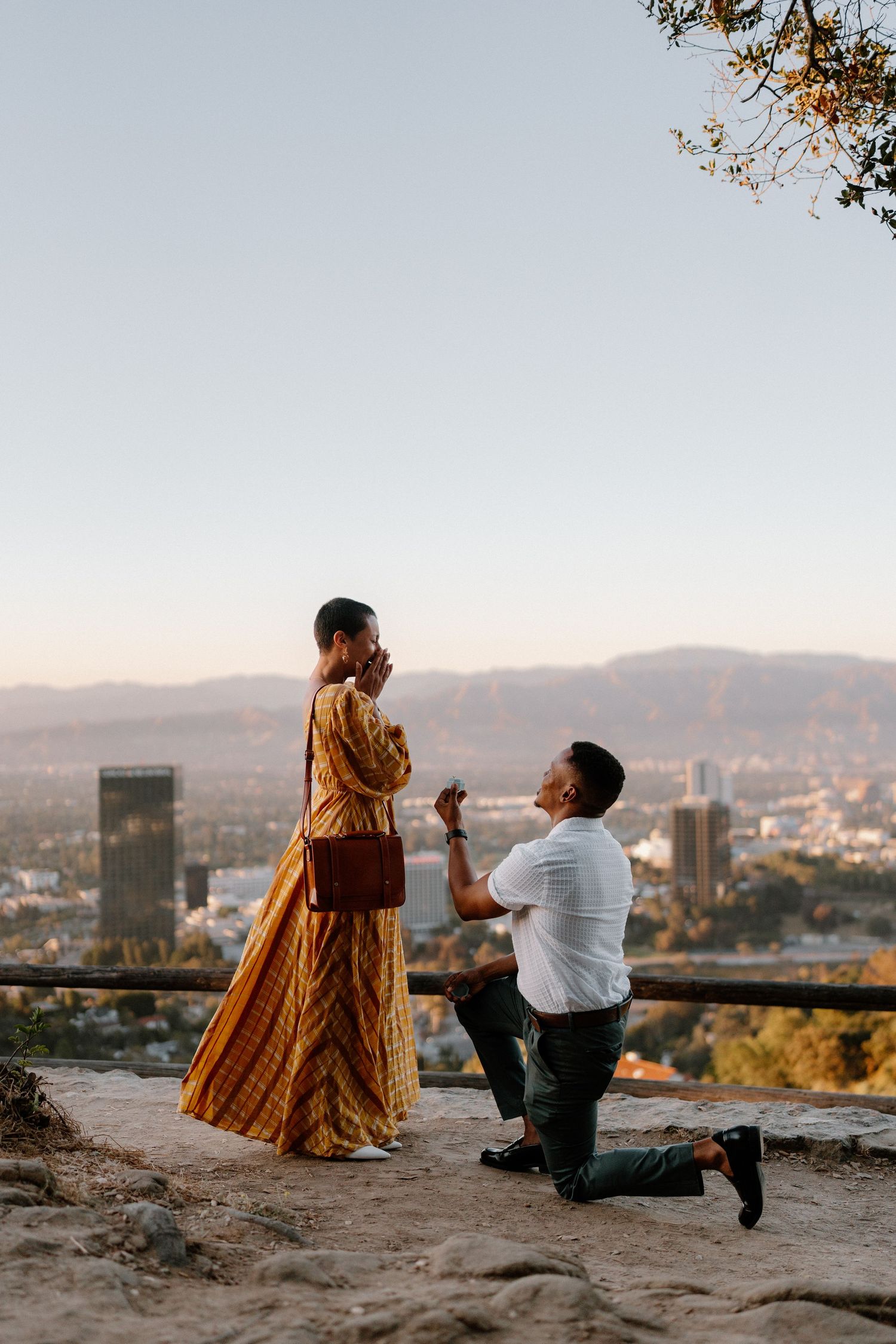 Man proposes on one knee to woman in golden dress at scenic LA overlook during sunset.