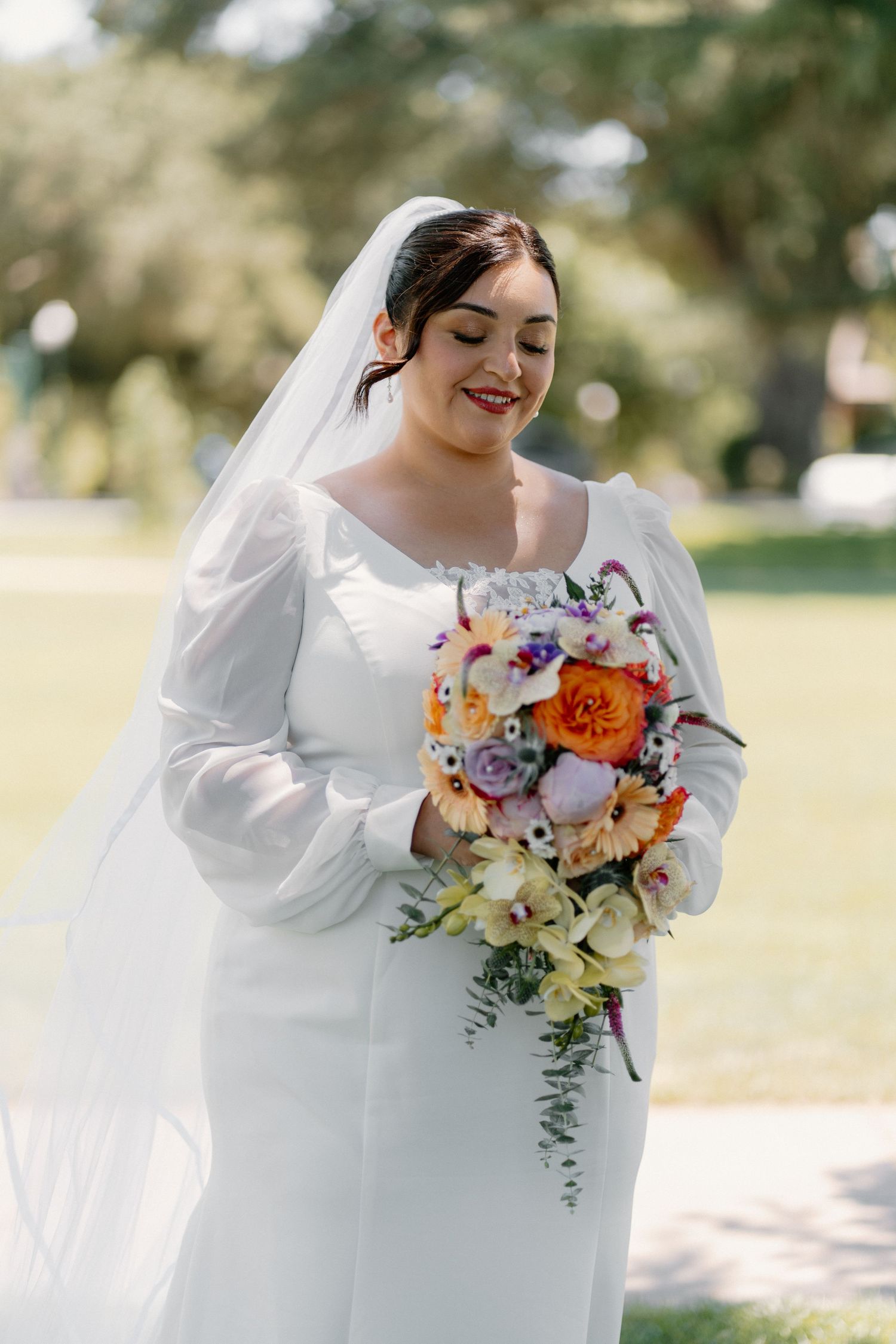 Bride in white gown with veil smiling down at vibrant orange and purple wedding bouquet outdoors.