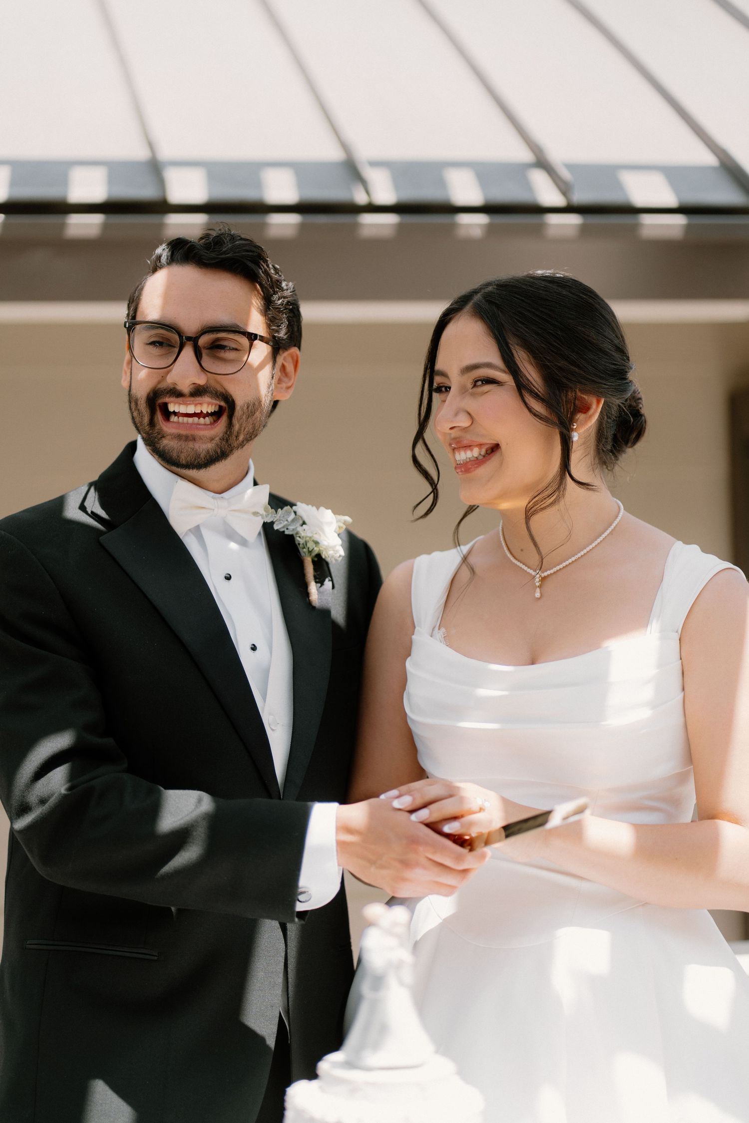 Smiling bride and groom in black tuxedo cutting wedding cake together in sunny outdoor setting.