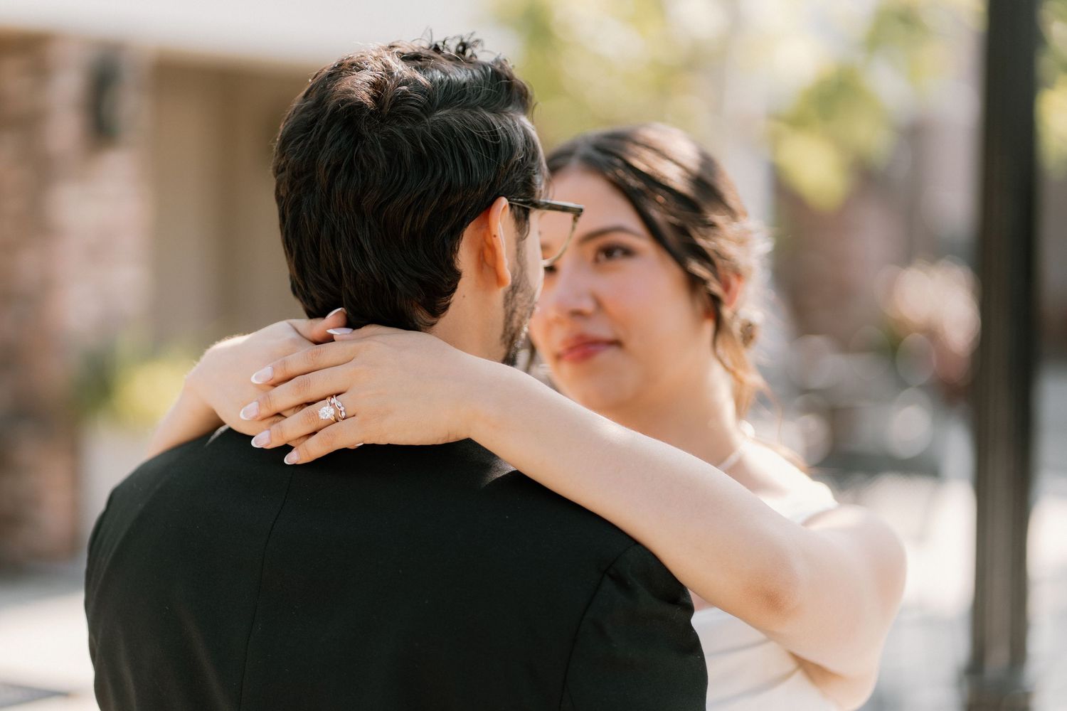 Bride with engagement ring embraces groom outdoors in warm sunlight during romantic wedding photo session.