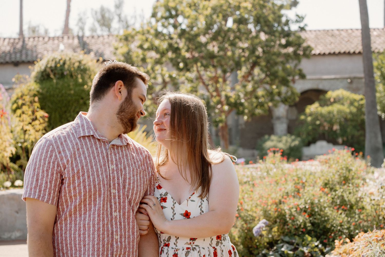 Couple smiling at each other in a sunny garden, woman in floral dress and man in striped shirt.
