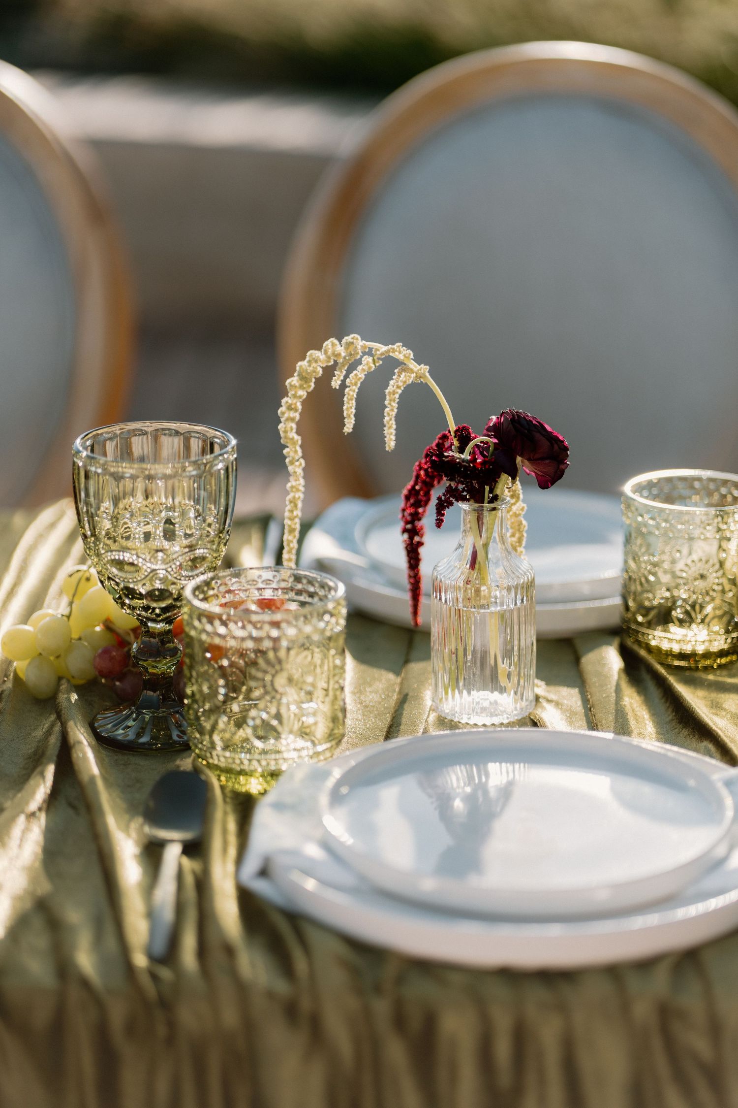 Elegant outdoor wedding table setting with gold glassware, white plates, dried florals, and grapes.