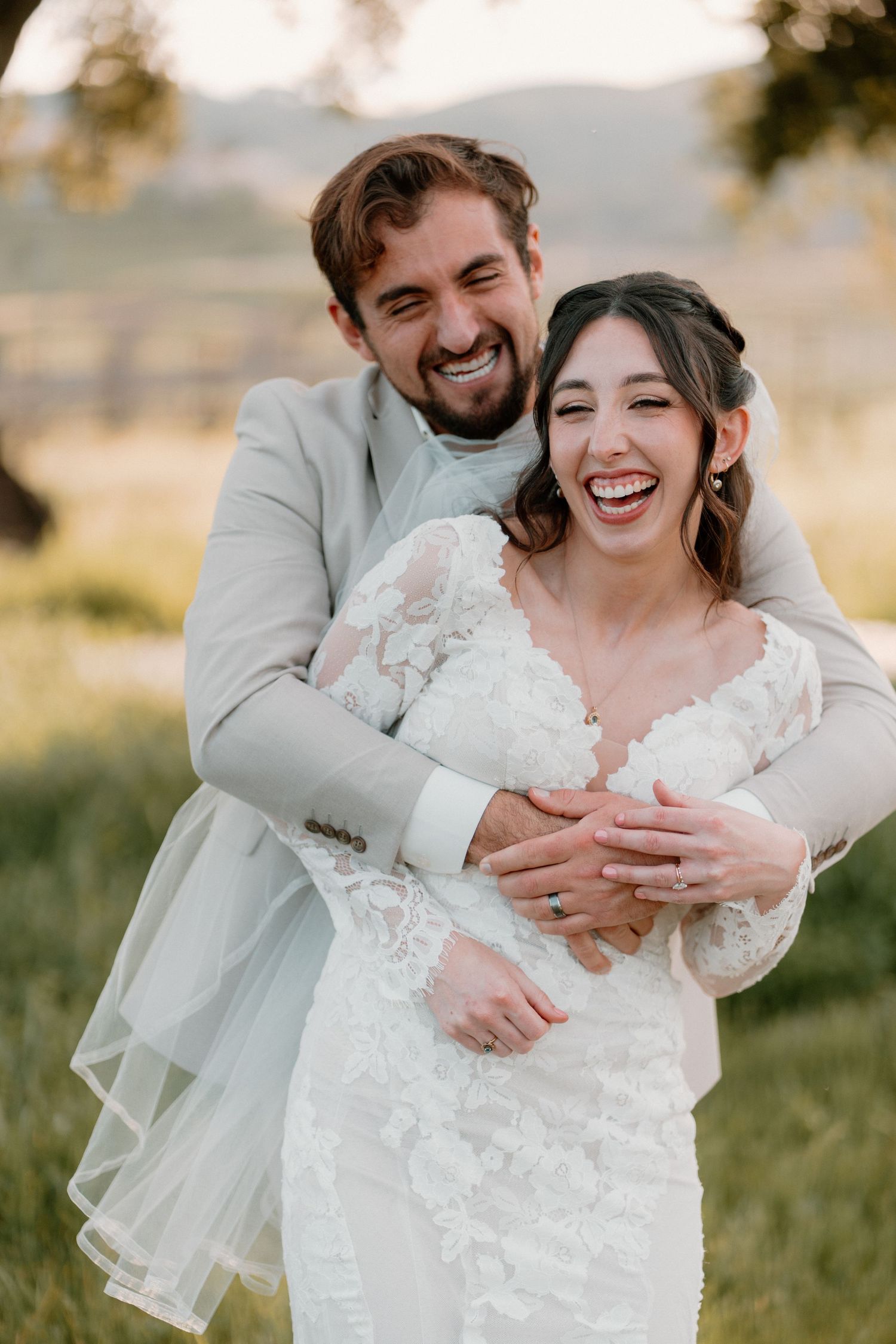 Joyful bride and groom laugh together as he hugs her from behind in a sunny outdoor wedding photo.