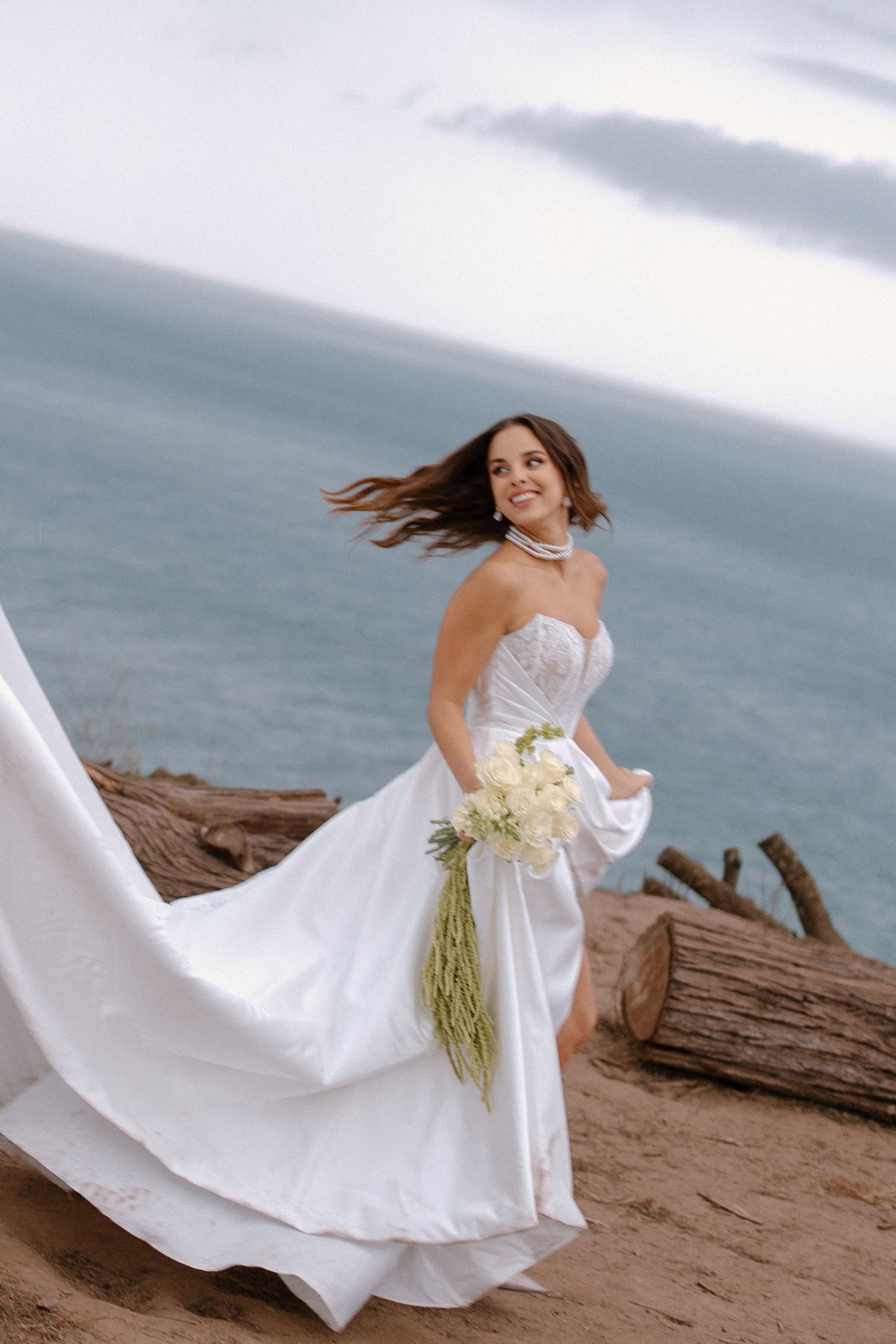 Joyful bride in flowing white strapless gown with pearl necklace poses on sandy cliff with bouquet, ocean in background.