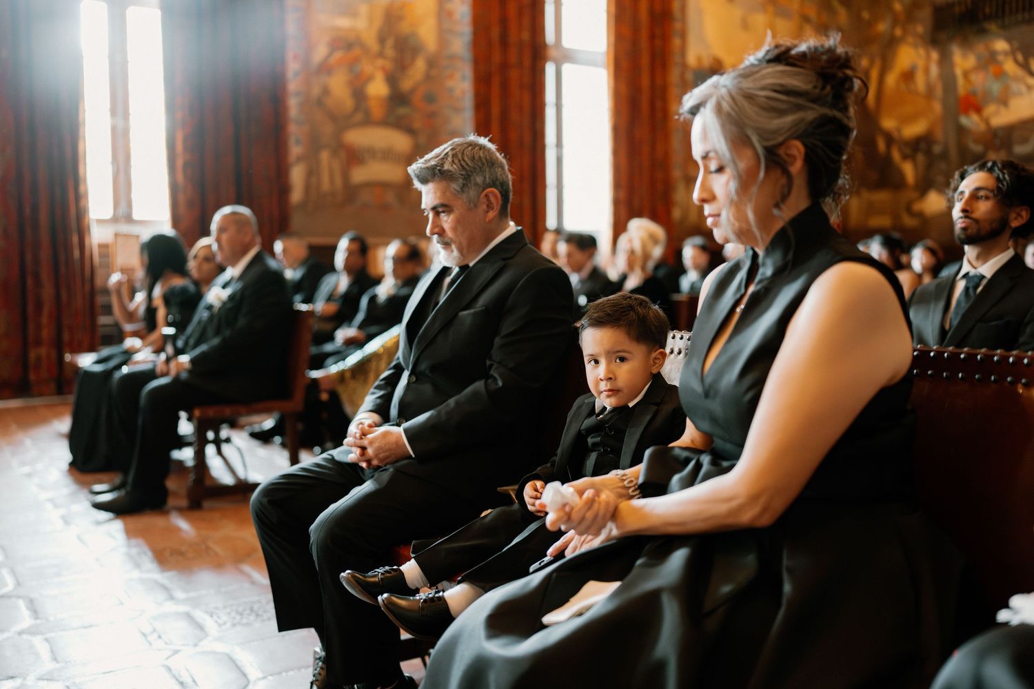 Wedding guests in formal black attire seated in ornate venue including a young boy in suit.