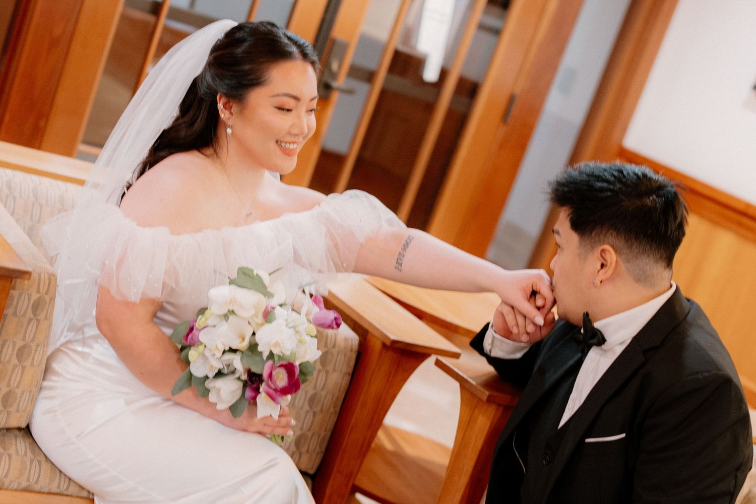 Smiling bride holding bouquet while groom kisses her hand tenderly inside a wedding venue.