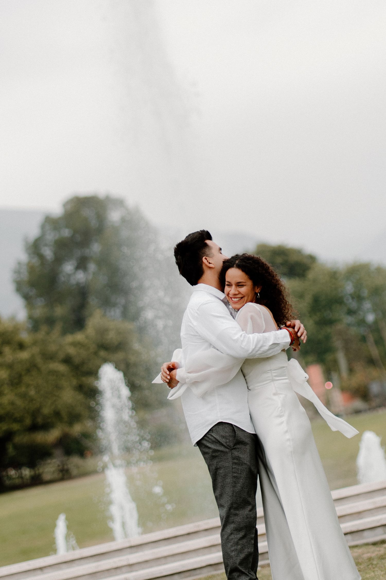 Happy couple embracing and laughing in front of a large outdoor fountain during engagement photos.