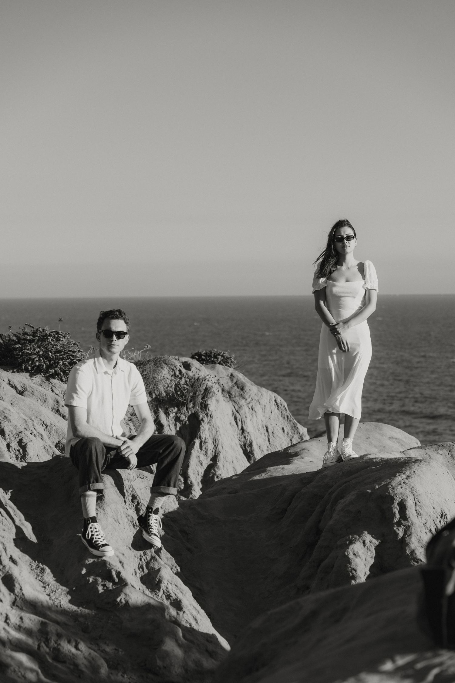 Black and white photo of a couple posing on coastal rocks overlooking the ocean at sunset.