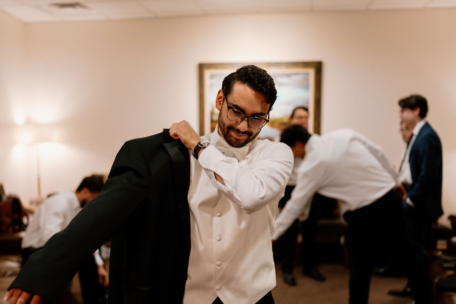 Smiling groom putting on tuxedo jacket with groomsmen getting ready in bright dressing room.