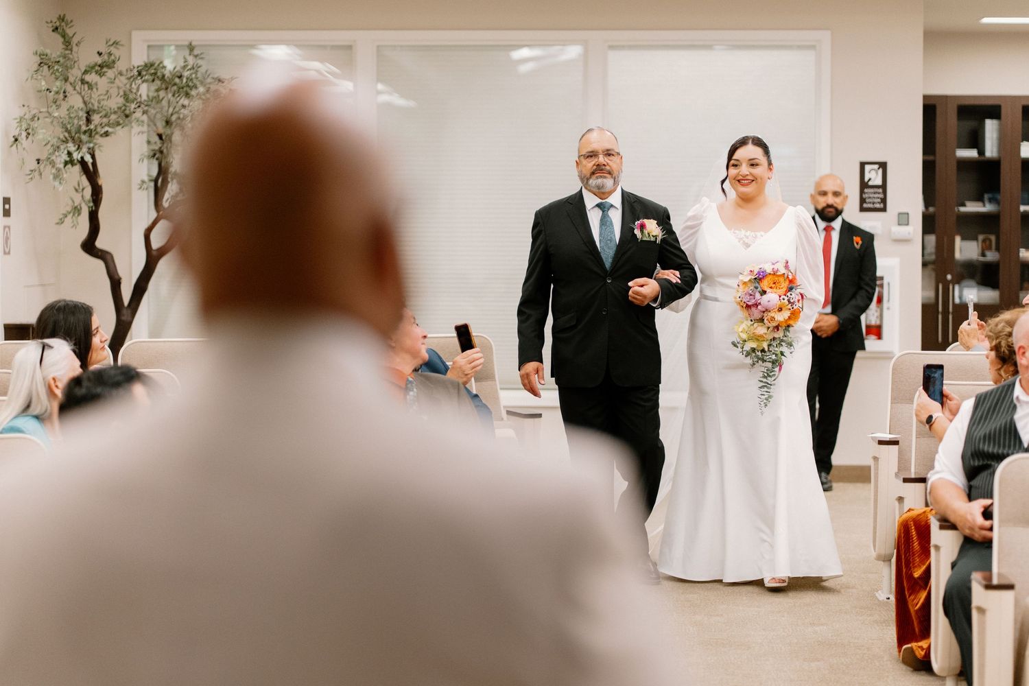 Bride walks down the aisle with her father at an indoor wedding ceremony, holding a colorful bouquet.