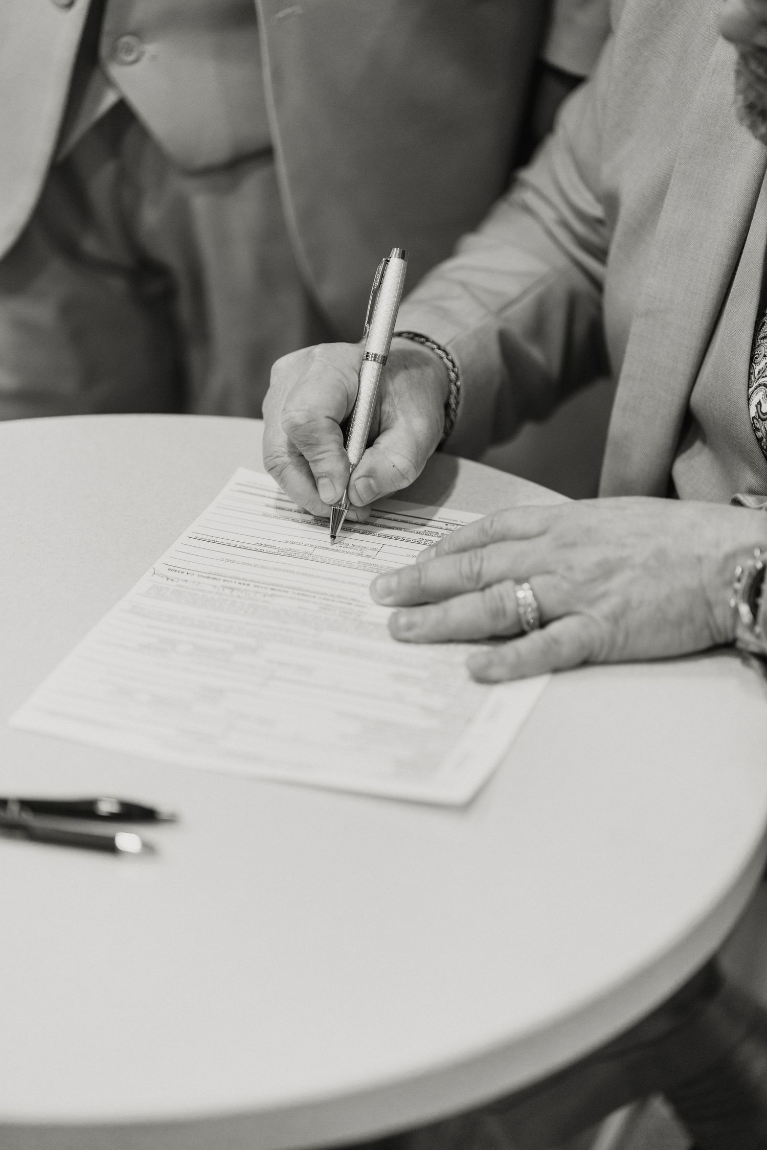 Black and white close-up of hands signing official wedding marriage license document at white table.