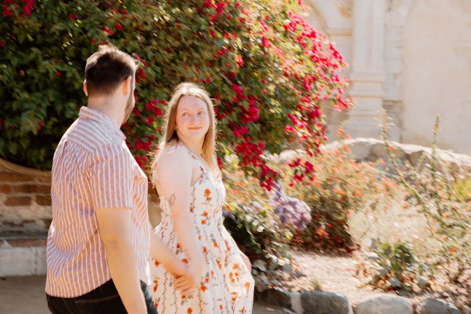 Couple holding hands outdoors near red bougainvillea flowers, woman in floral dress smiling at man in striped shirt.