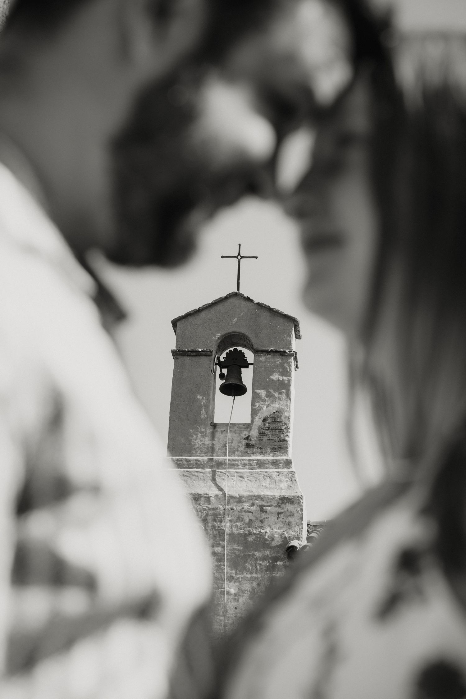 A blurred couple frames a black and white shot of a historic bell tower with a cross on top.