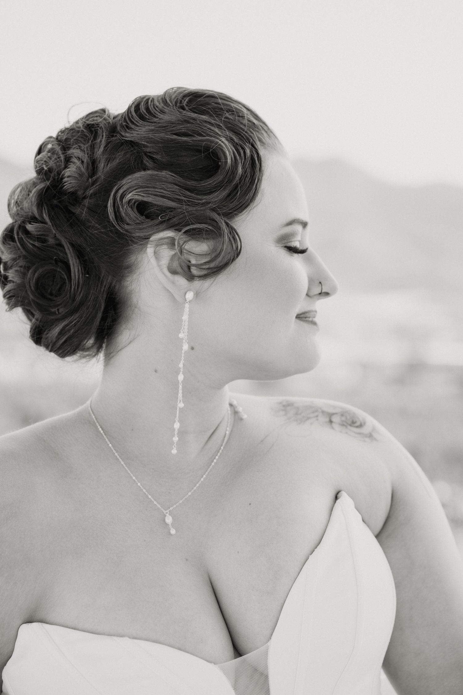 Black and white portrait of bride with elegant updo, pearl drop earrings, and floral shoulder tattoo.