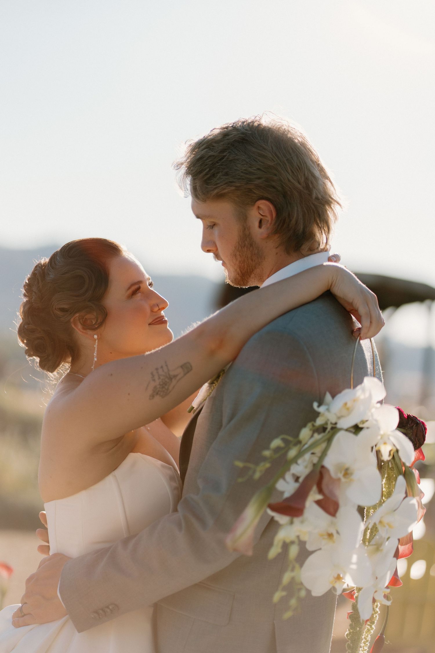 Bride embracing groom at golden hour holding white orchid bouquet outdoors.