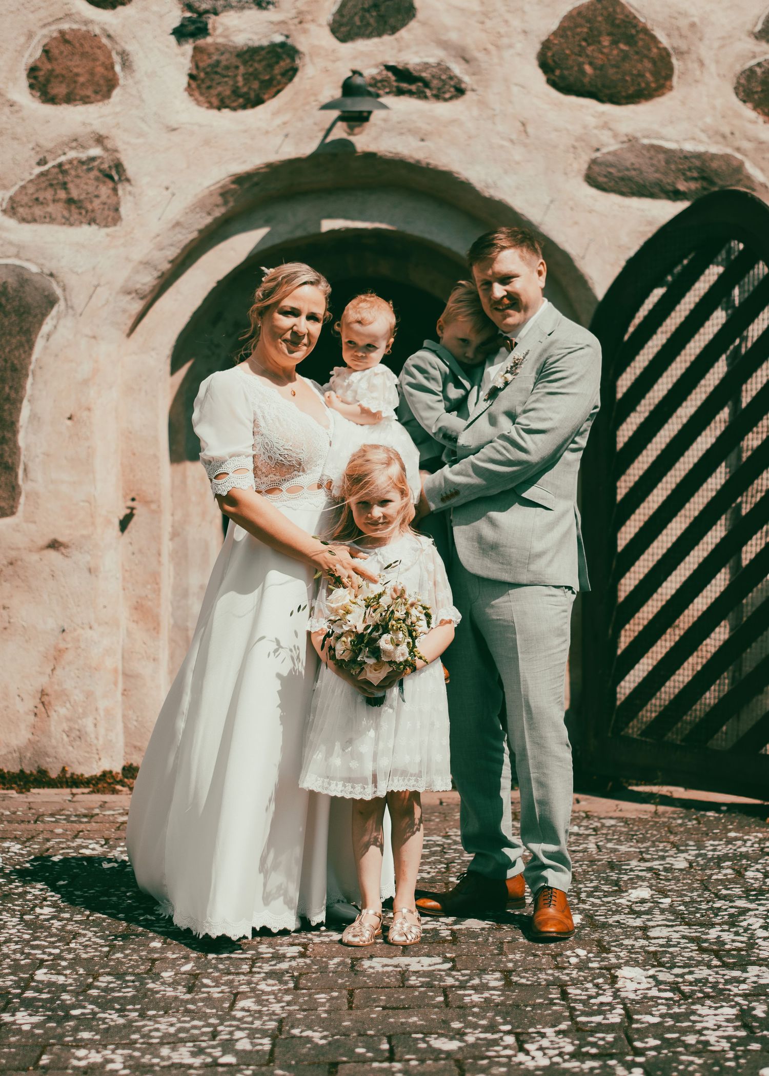 Hochzeitspaar mit Kindern vor einer rustikalen Steinmauer