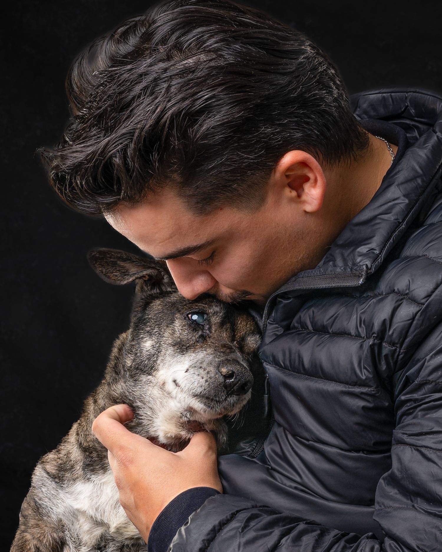 A young man in a navy puffer jacket tenderly kisses his brindle dog against a dark background.