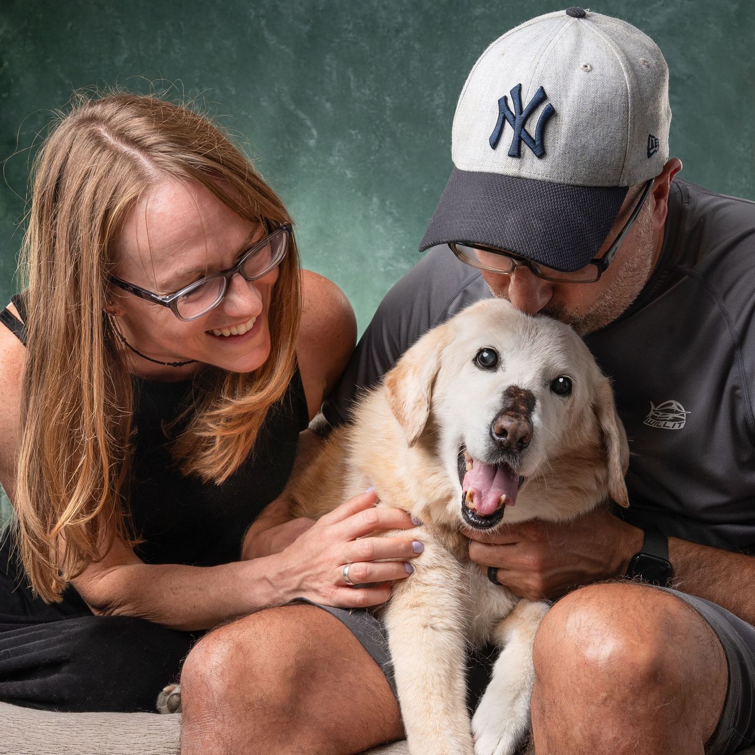 A smiling couple with glasses and NY Yankees cap cuddles their happy golden Labrador dog against a green backdrop.