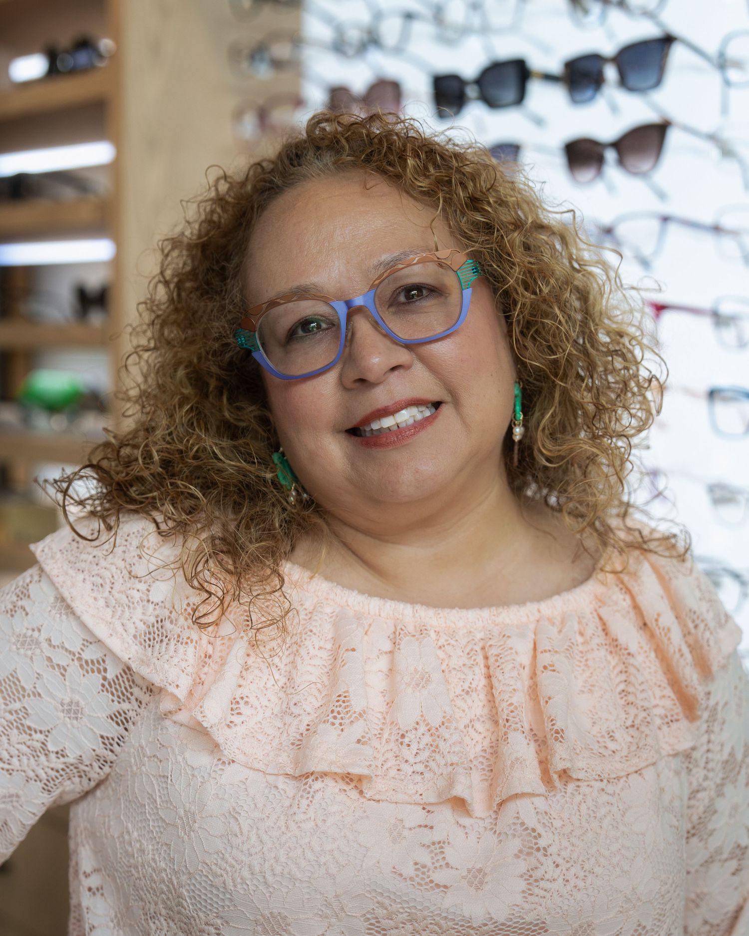 Woman with curly hair wearing blue glasses and pink lace top smiles in optical store with eyeglass frames displayed.