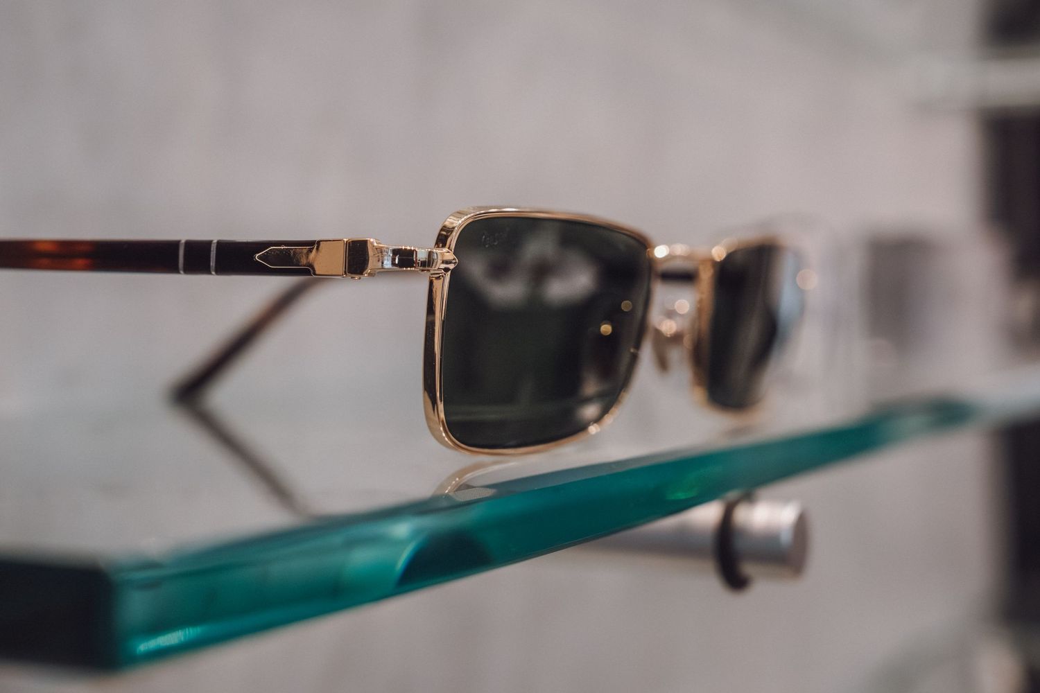 Gold-framed square sunglasses with dark lenses displayed on a glass shelf, reflecting elegantly on the surface.