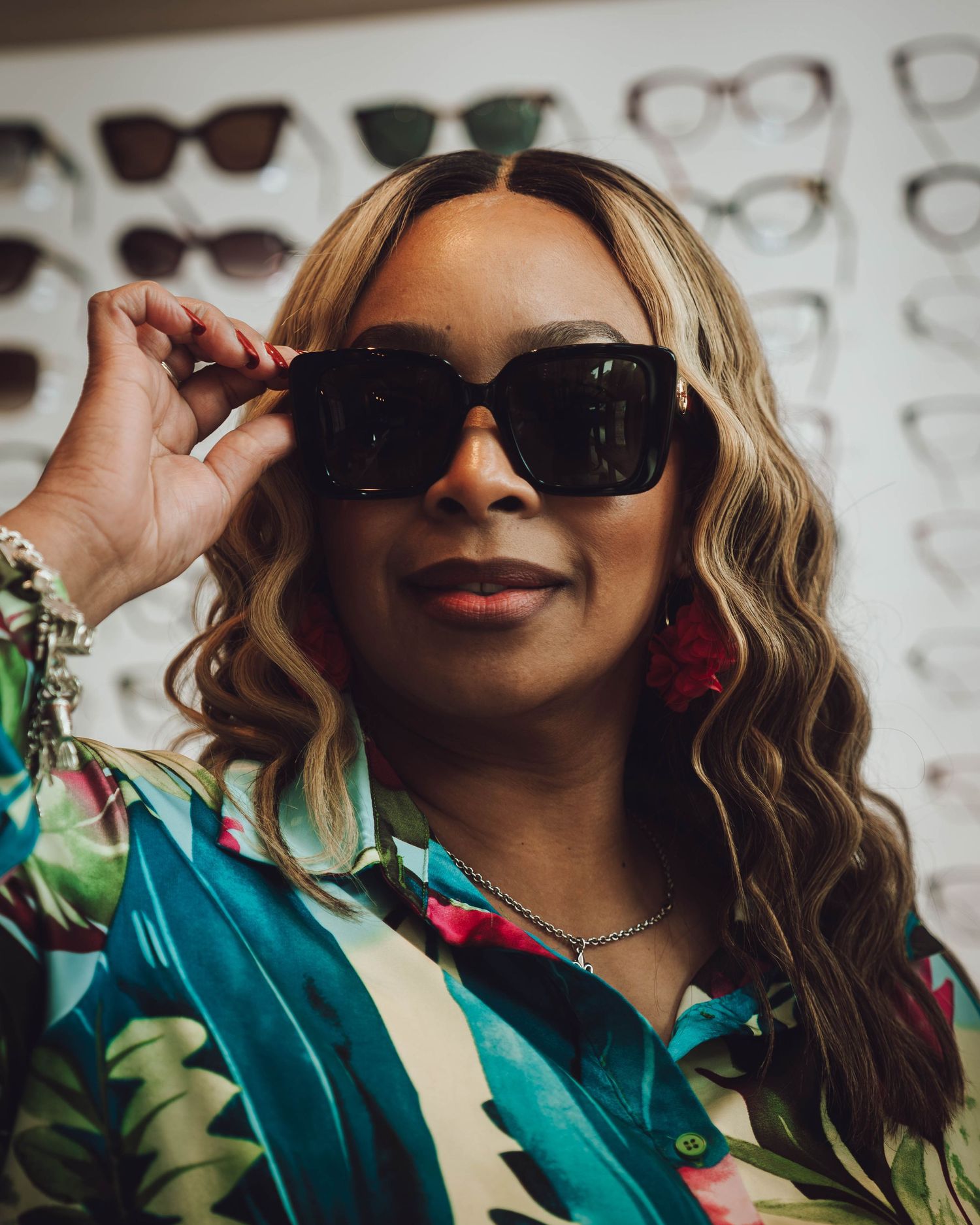 Woman trying on large black square sunglasses in an eyewear store, wearing a floral top and flower earrings.