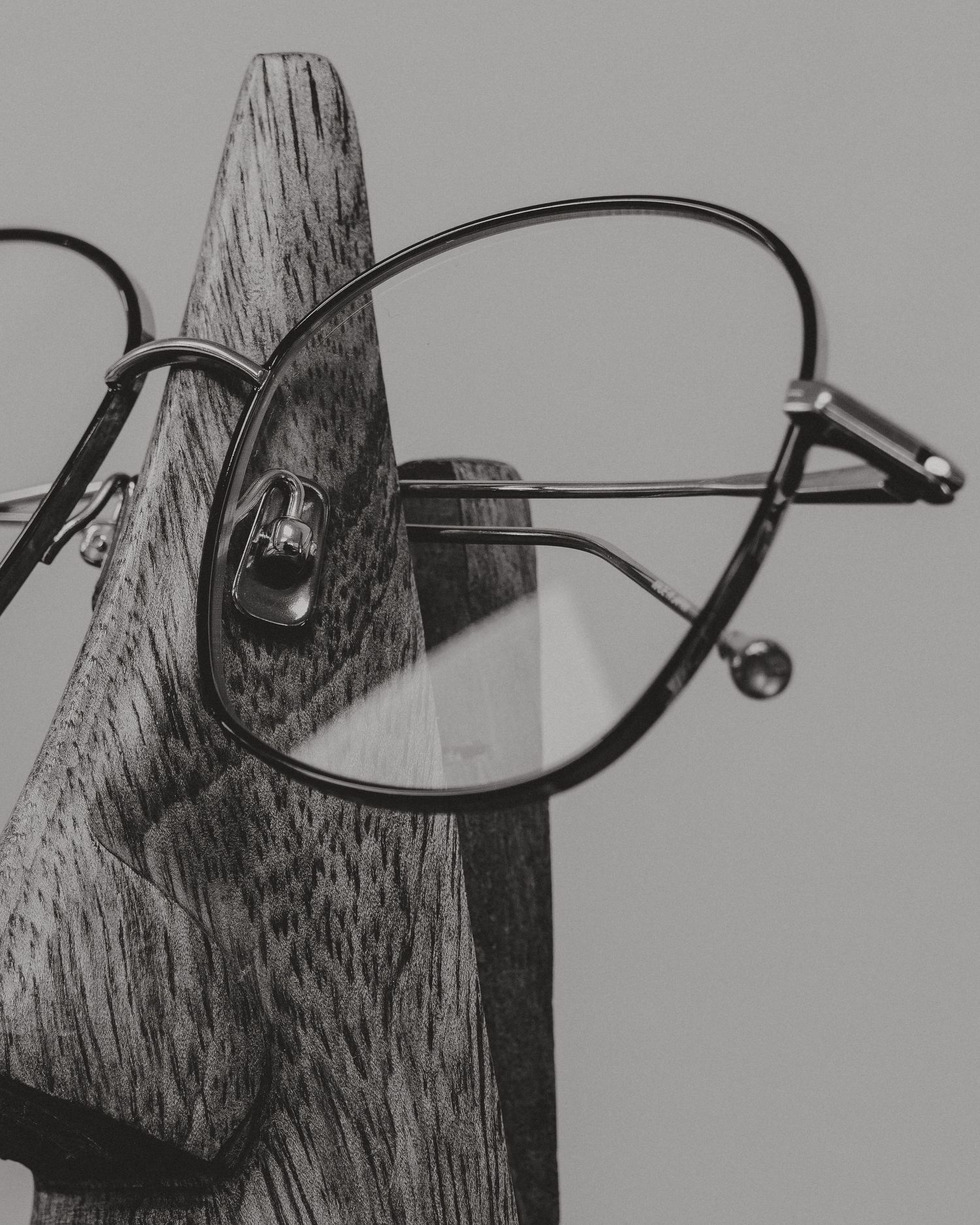 Black and white photo of round eyeglasses resting on a wooden nose-shaped stand against a gray background.