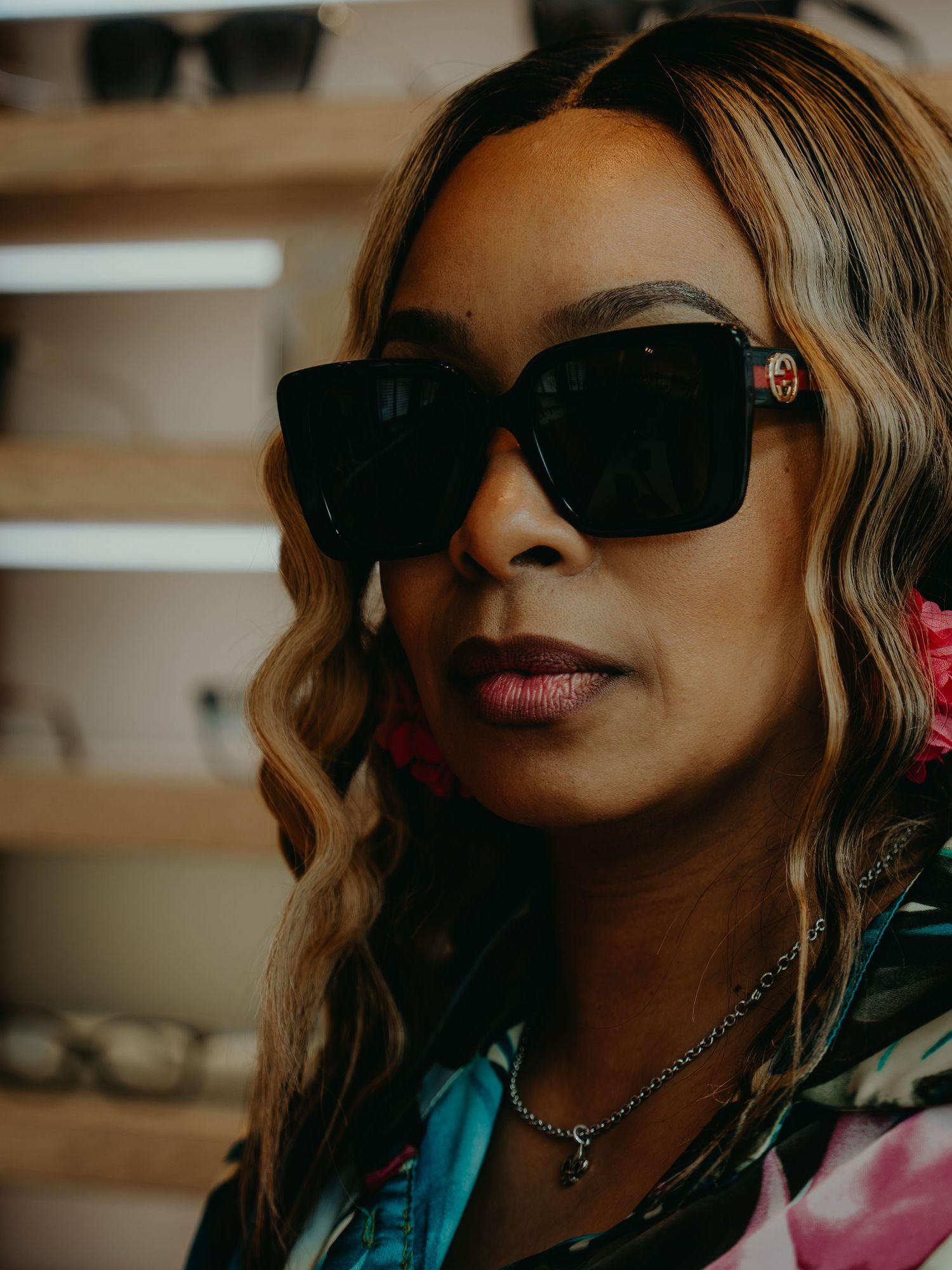 Woman wearing large black Gucci sunglasses with wavy blonde hair and a colorful jacket in an eyewear store.