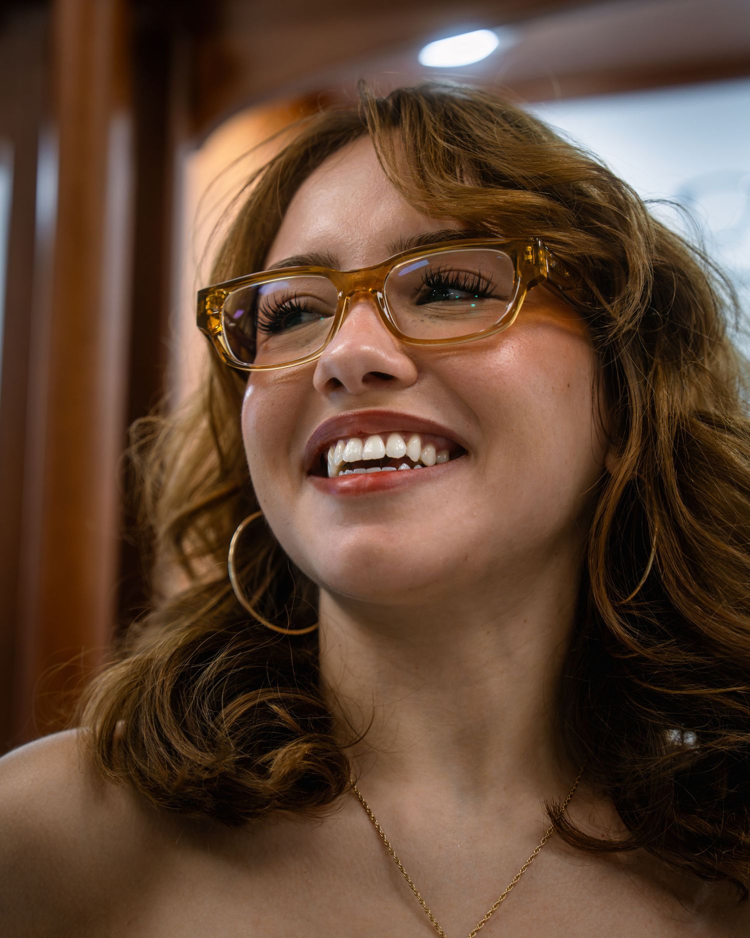 Smiling woman with auburn hair, amber glasses, hoop earrings, and a gold necklace laughing joyfully.