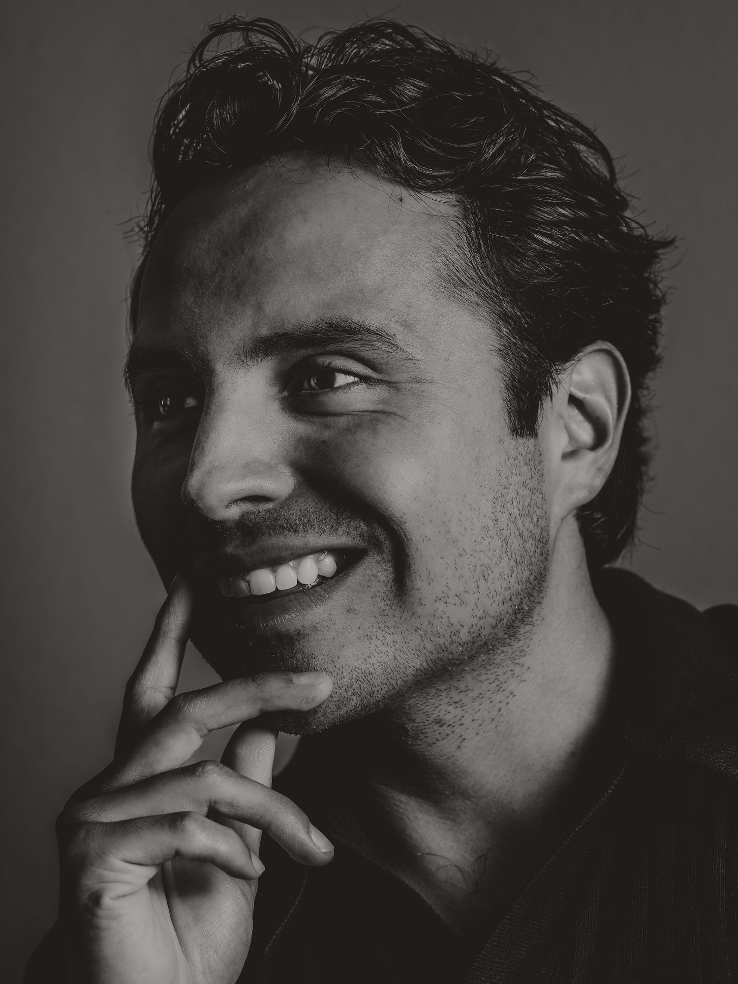 Black and white portrait of a smiling young man with stubble, hand on chin, looking upward thoughtfully.