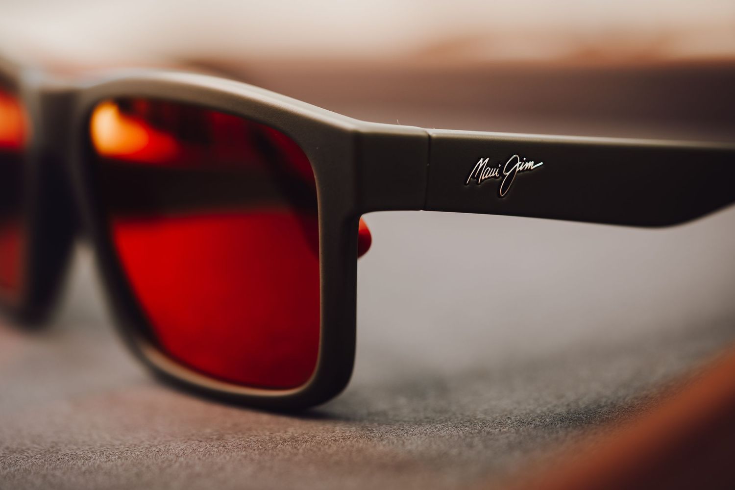 Maui Jim sunglasses with red polarized lenses and matte black frames resting on a gray surface.