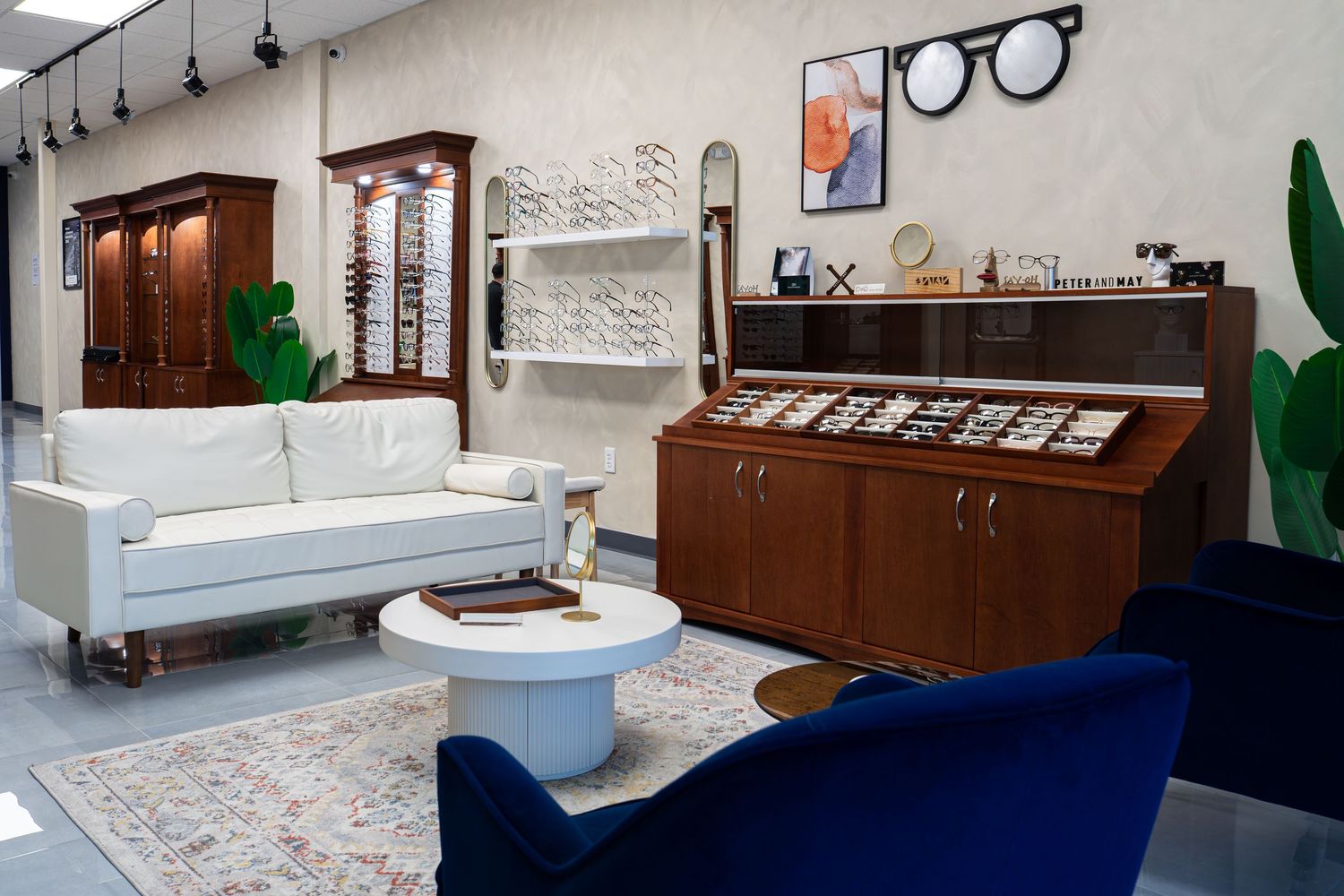 Modern optical store showroom with white sofa, navy chairs, wood display cabinet, and eyeglass frames on wall-mounted racks.