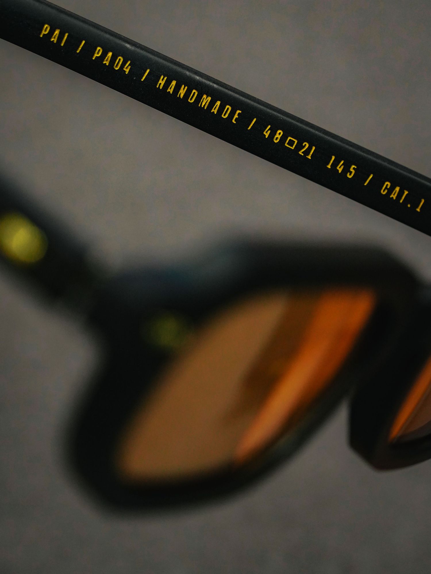 Close-up of PAI PA04 handmade sunglasses arm with gold text, featuring amber mirrored lenses on dark background.