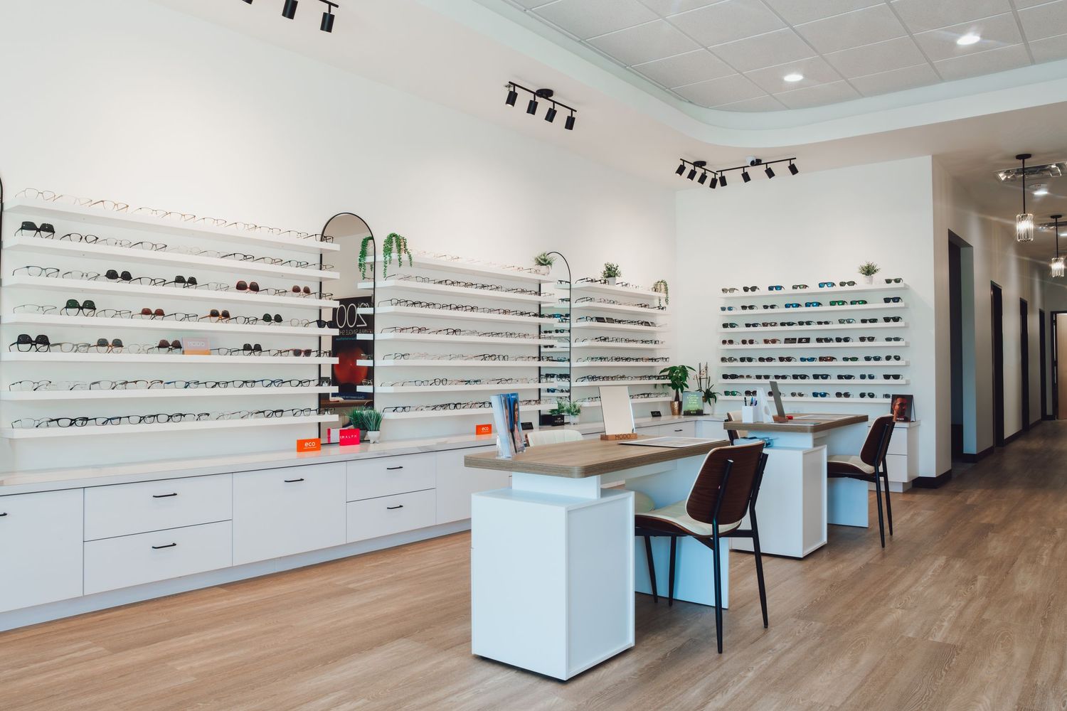 Modern optical store interior with white display shelves of eyeglasses, wooden consultation desks, and chairs on hardwood floors.