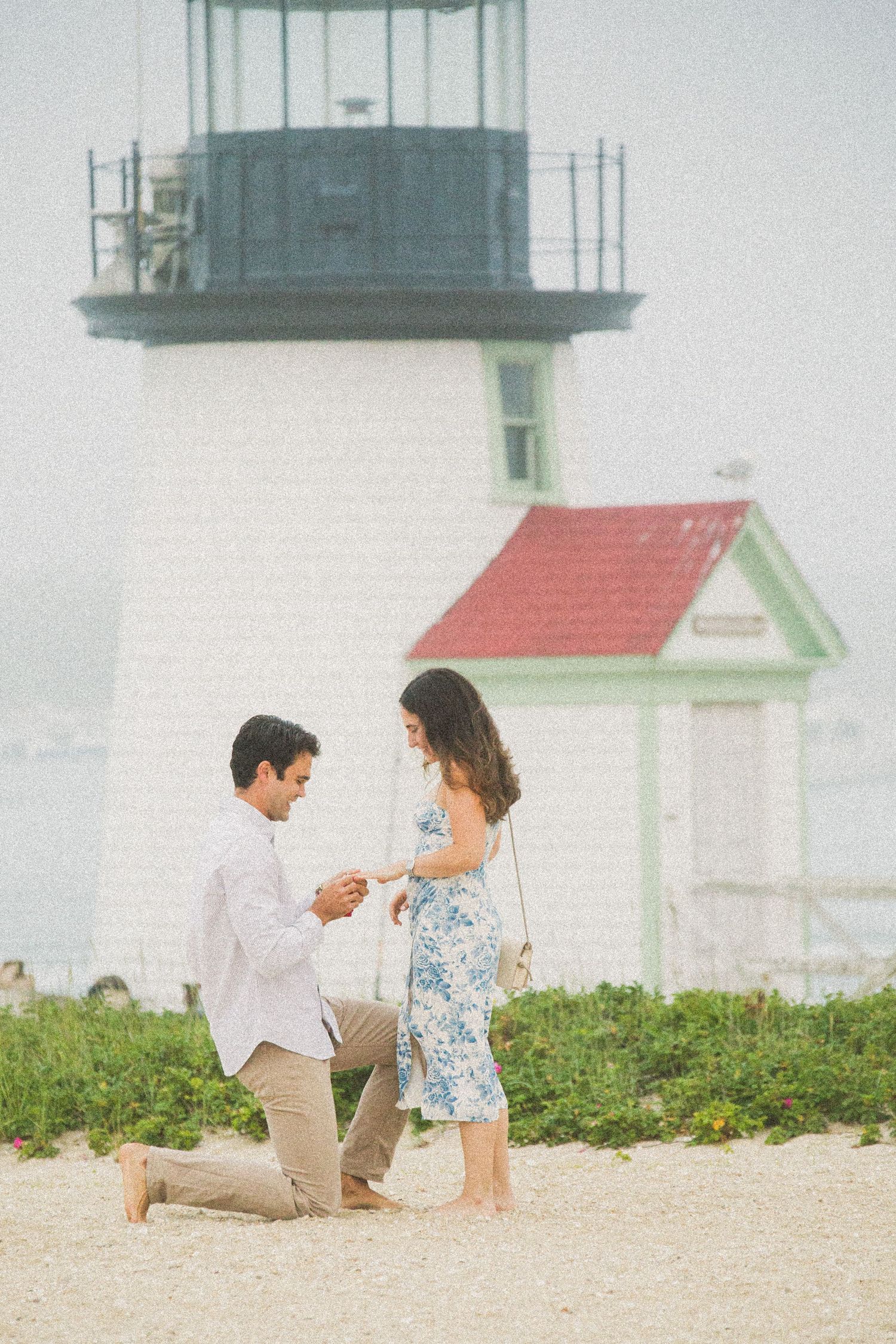 Man proposes to woman on sandy beach with historic white lighthouse and red roof building in background.