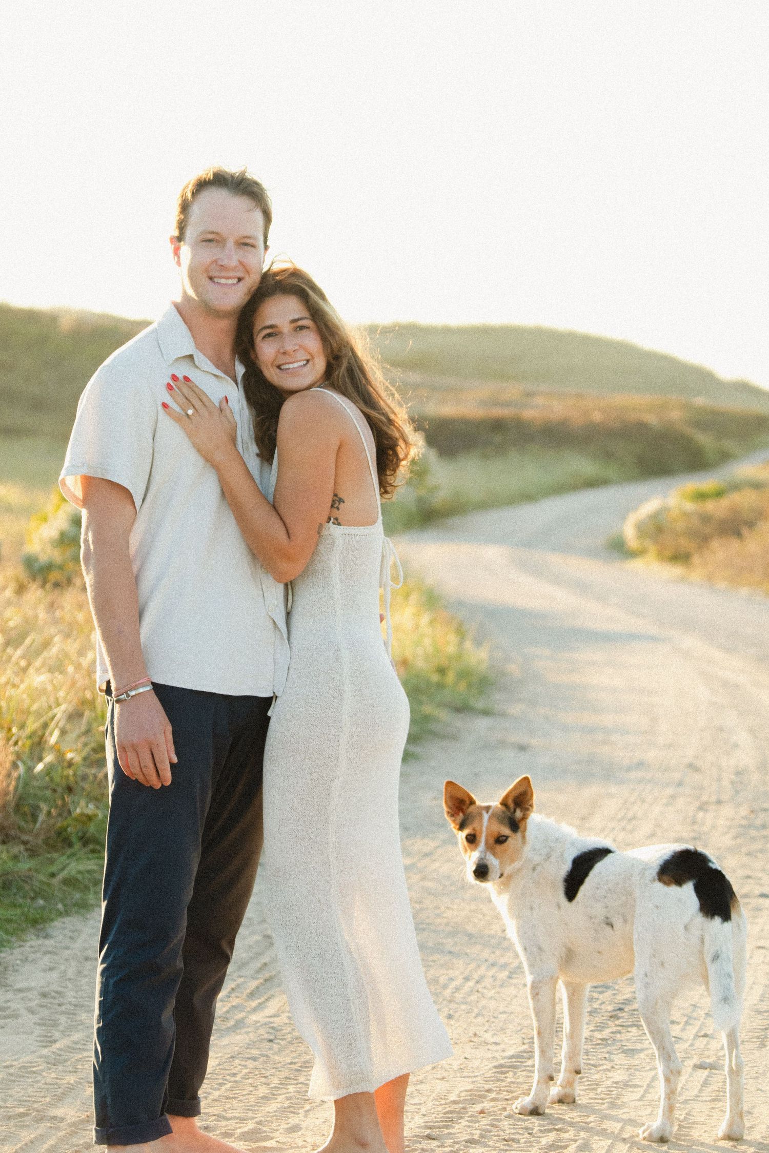 Couple embracing on dirt path at sunset with two dogs walking beside them.