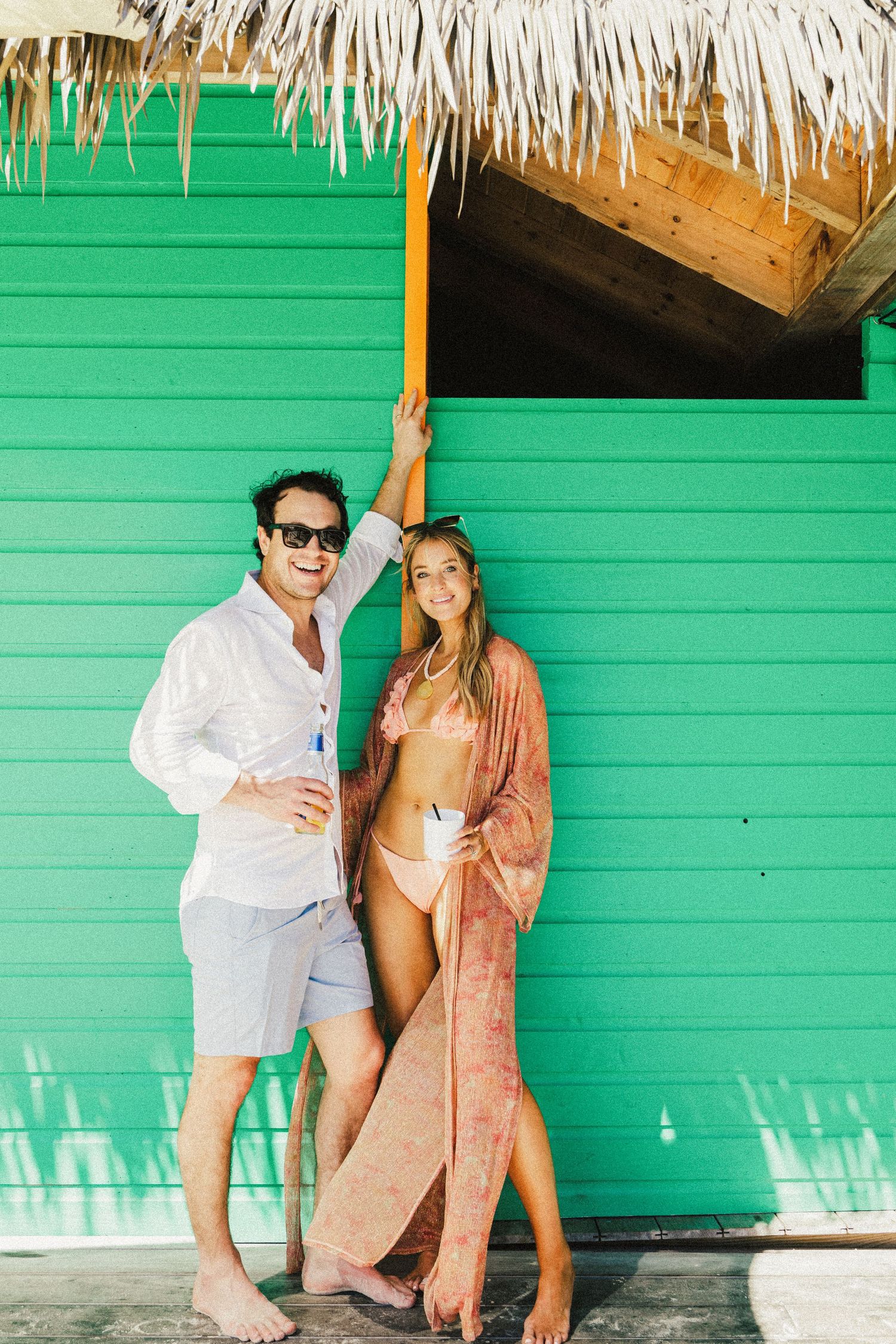 Couple posing against a bright green wall with thatched roof overhang at a tropical resort.