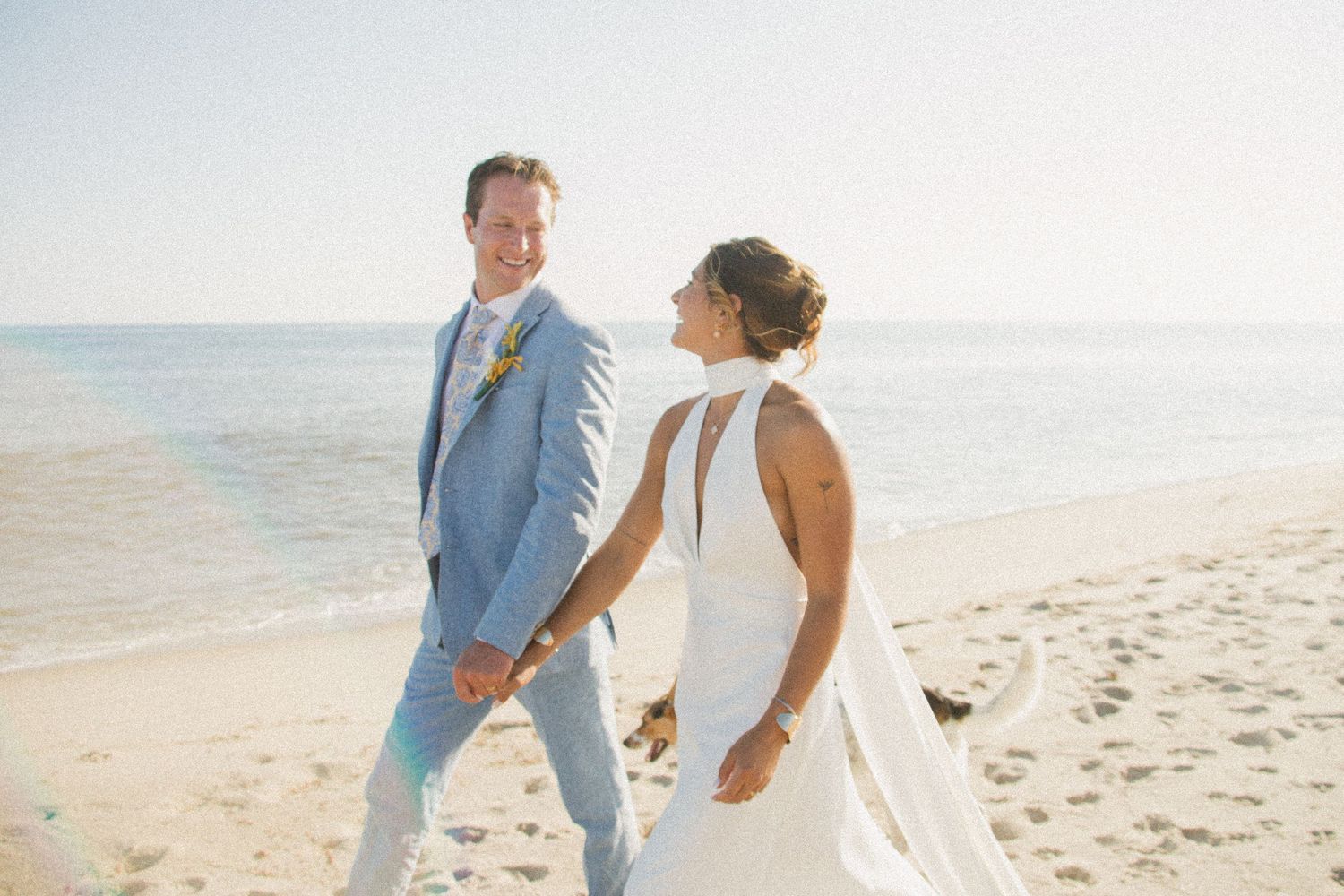 Sun-drenched beach wedding portrait featuring elegant bridal gown and light blue suit.