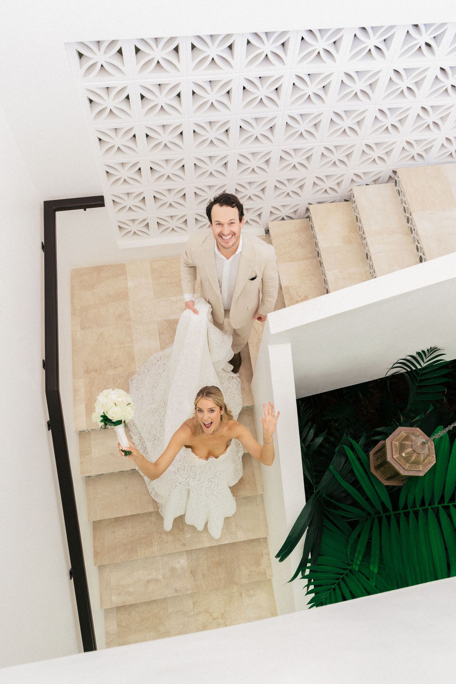 A joyful wedding couple poses on elegant stone stairs with a decorative ceiling pattern.